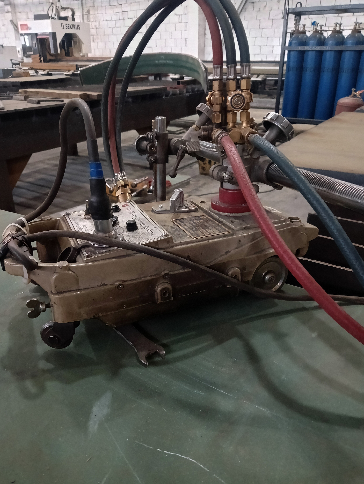 An industrial scene is depicted, focusing on a portable oxy-fuel cutting machine situated on a green surface, likely a workbench or table. The machine, a beige metallic unit with wheels, is connected via thick, multi-colored hoses to what appear to be gas cylinders, visible in the background. One hose is red, another is dark gray, and a third is teal. The machine itself has various controls and a metallic plate with text, indicating it's a "FG-50 Gas Cutting Machine". In the background, a tall rack holds several blue gas cylinders. To the left, a large white and black machine with the "SEKIRUS" logo is partially visible. The environment suggests a workshop or factory setting, with other industrial equipment and materials scattered around. The lighting is artificial, creating shadows on the floor. There are no people in the frame.