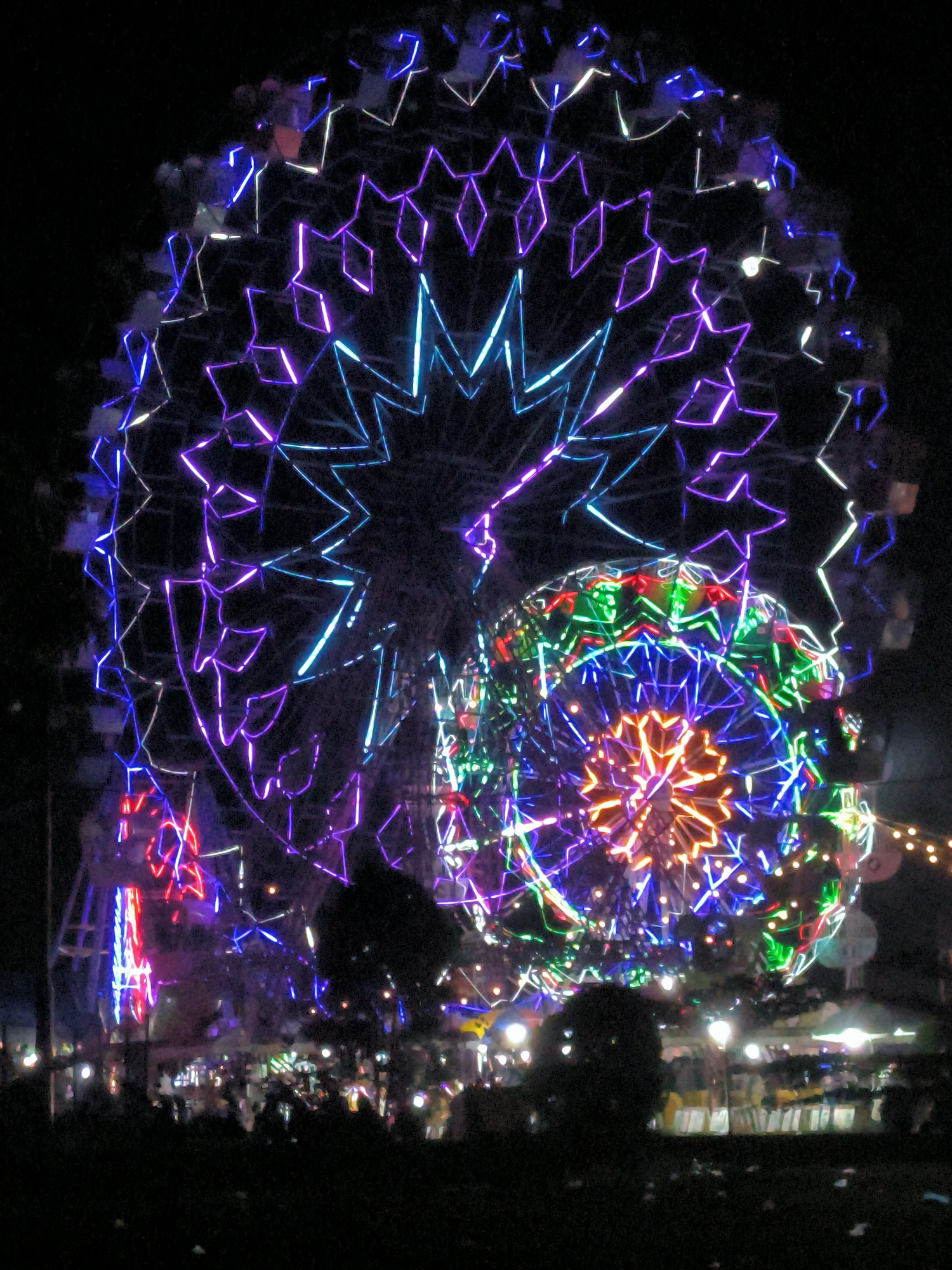 At night in Titibaro, Indonesia, two illuminated Ferris wheels dominate the scene. The larger wheel is predominantly blue and purple, displaying geometric light patterns. A smaller, multi-colored Ferris wheel sits in front of the larger one. Below the Ferris wheels, a crowd is visible, with numerous stalls or vendors and illuminated lights interspersed among the crowd. Dark silhouettes of trees or bushes frame the foreground. The sky is dark, and the overall scene suggests a festive or celebratory event.