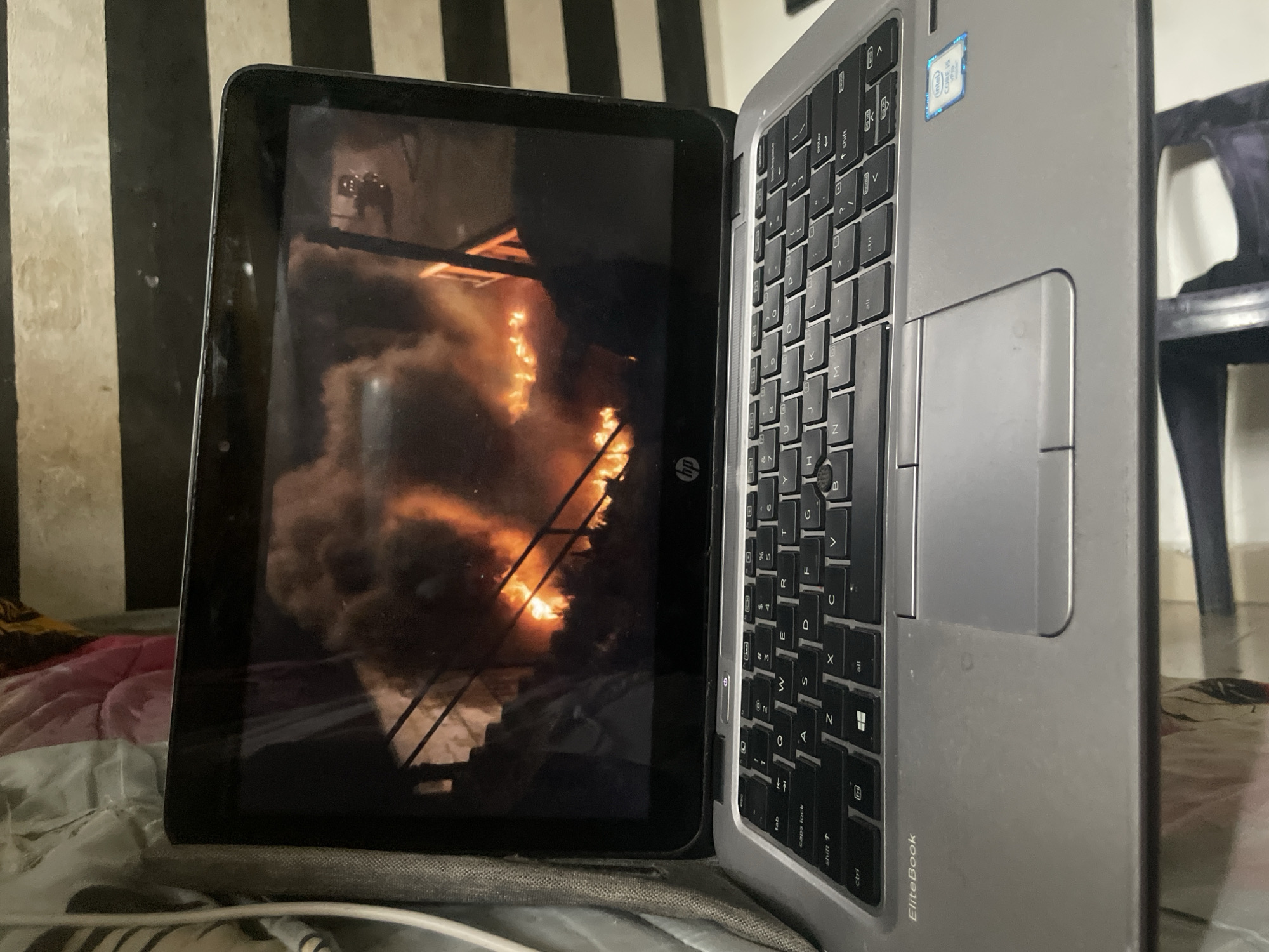 The image shows a laptop with a screen displaying a fire. The laptop is open and placed on a bed or couch. The screen of the laptop shows a large fire with smoke billowing upwards. The laptop is a silver HP EliteBook. The background of the image shows a striped wall, possibly wallpaper or painted stripes. The scene is indoors, and the lighting suggests it might be nighttime or a dimly lit room. There is no indication of the location in the city of Ihiagwa, Nigeria.