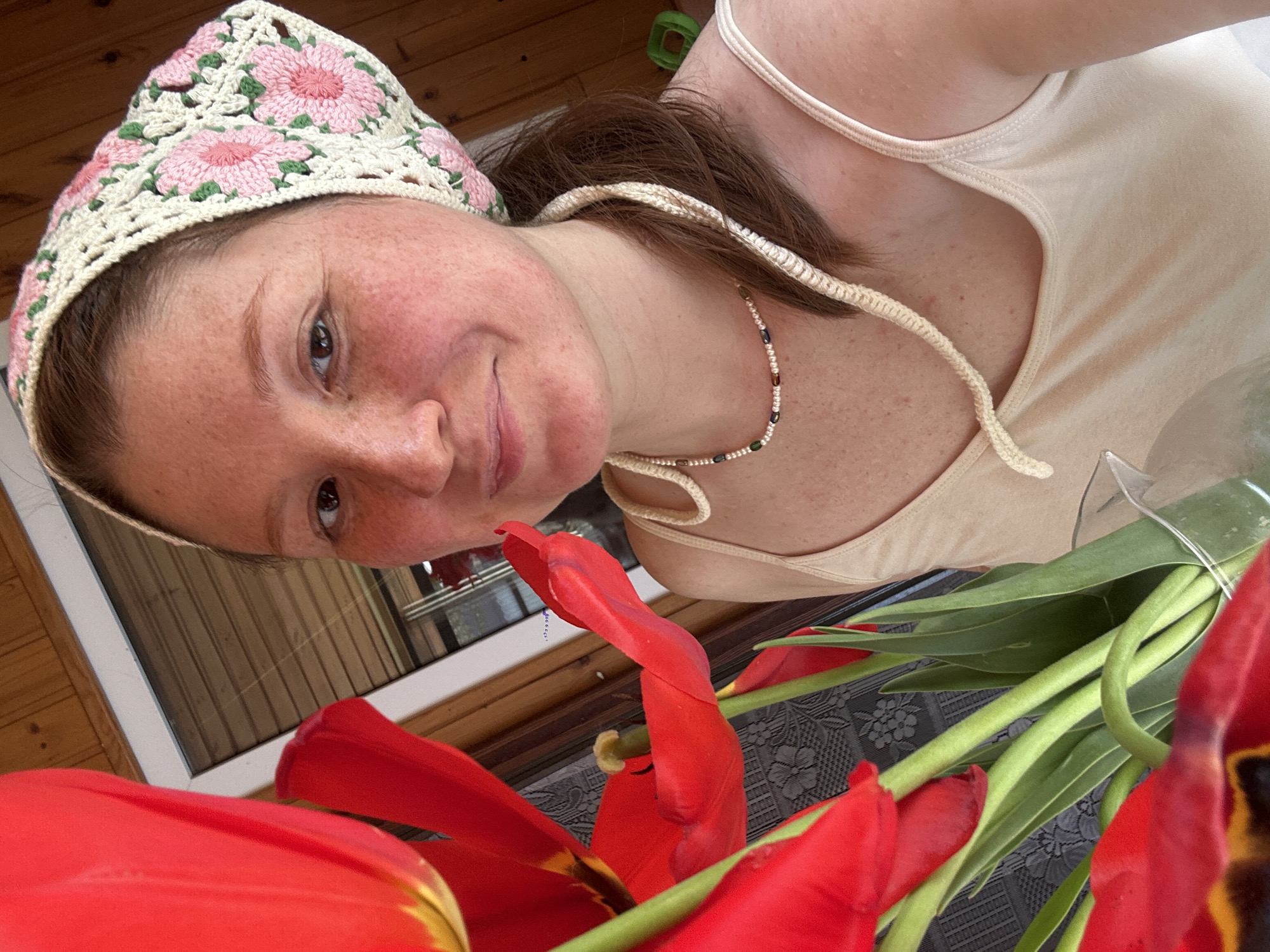 A woman with reddish-brown hair and freckles is visible in a close-up, angled shot. She is wearing a light beige tank top and a crocheted head covering with pink floral designs. A beaded necklace adorns her neck. Below her face and torso, a bouquet of red tulips is positioned. The flowers are tied with a clear plastic band, and their green stems are visible. The background features wooden paneling and a window frame. The location is suggested to be Nizhny Novgorod, Russia.