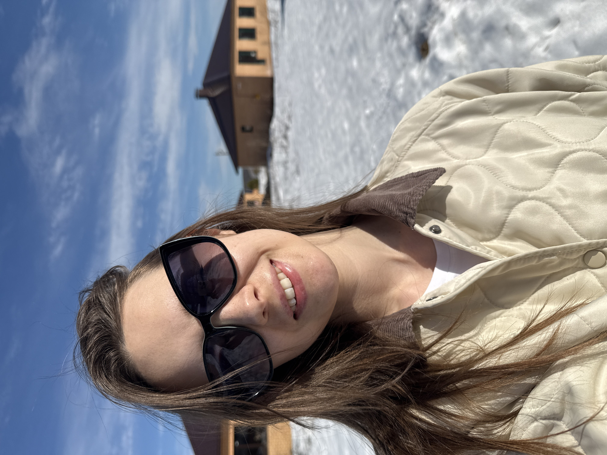A selfie photograph depicts an unidentified adult female outdoors in Chaikovsky, Russia, under bright daylight. The individual, positioned centrally, faces the camera with a visible smile, exposing her upper and lower teeth. She has medium-brown hair, with strands lifted by what appears to be wind, and wears dark-lensed, black-framed sunglasses. Her attire includes a light cream or beige quilted jacket with a brown corduroy collar and a white undershirt. The background features a snow-covered field, partially melted to reveal darker ground. Two detached, single-story residential buildings are visible in the mid-ground. The most prominent structure, on the right, is light brown with a dark roof, a central brick chimney, and multiple dark-framed windows. A second, similar structure is partially visible on the left. The sky overhead is clear blue with sparse, wispy white clouds.