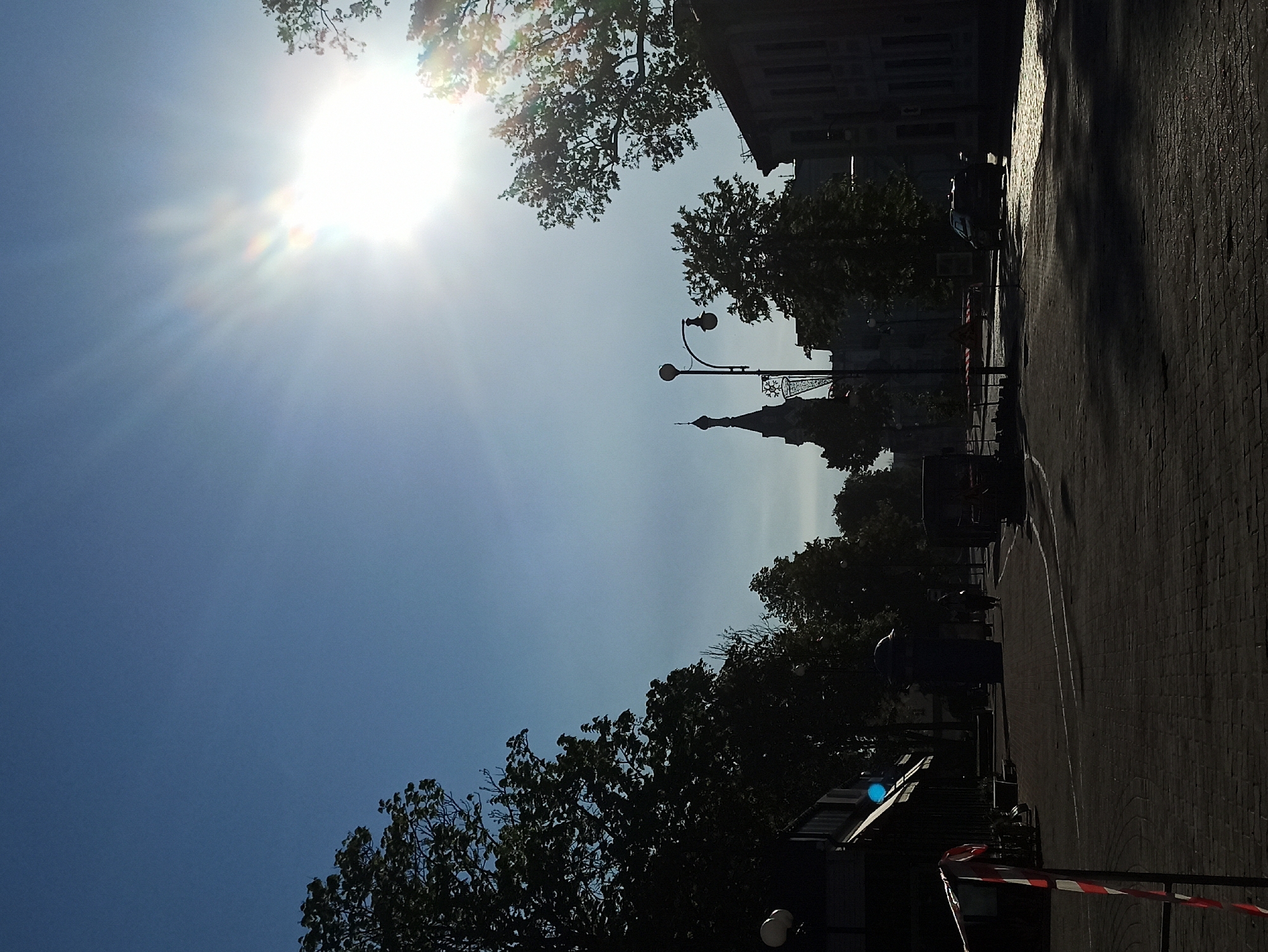 The image shows a street scene in Hrodna, Belarus, under bright sunlight. A spire-like structure is visible in the distance, possibly a church or cathedral. A row of lampposts lines the street. The street itself is paved with bricks and appears to be wet in sections, reflecting the sunlight. Trees are present on both sides of the street, casting shadows. A few indistinct figures of people can be seen in the middle ground. A building is situated on the right side of the street, partially obscured by trees. Red and white tape is present in the foreground, possibly indicating a restricted area. A vehicle is parked on the roadside further up the street. The sky is clear and blue.