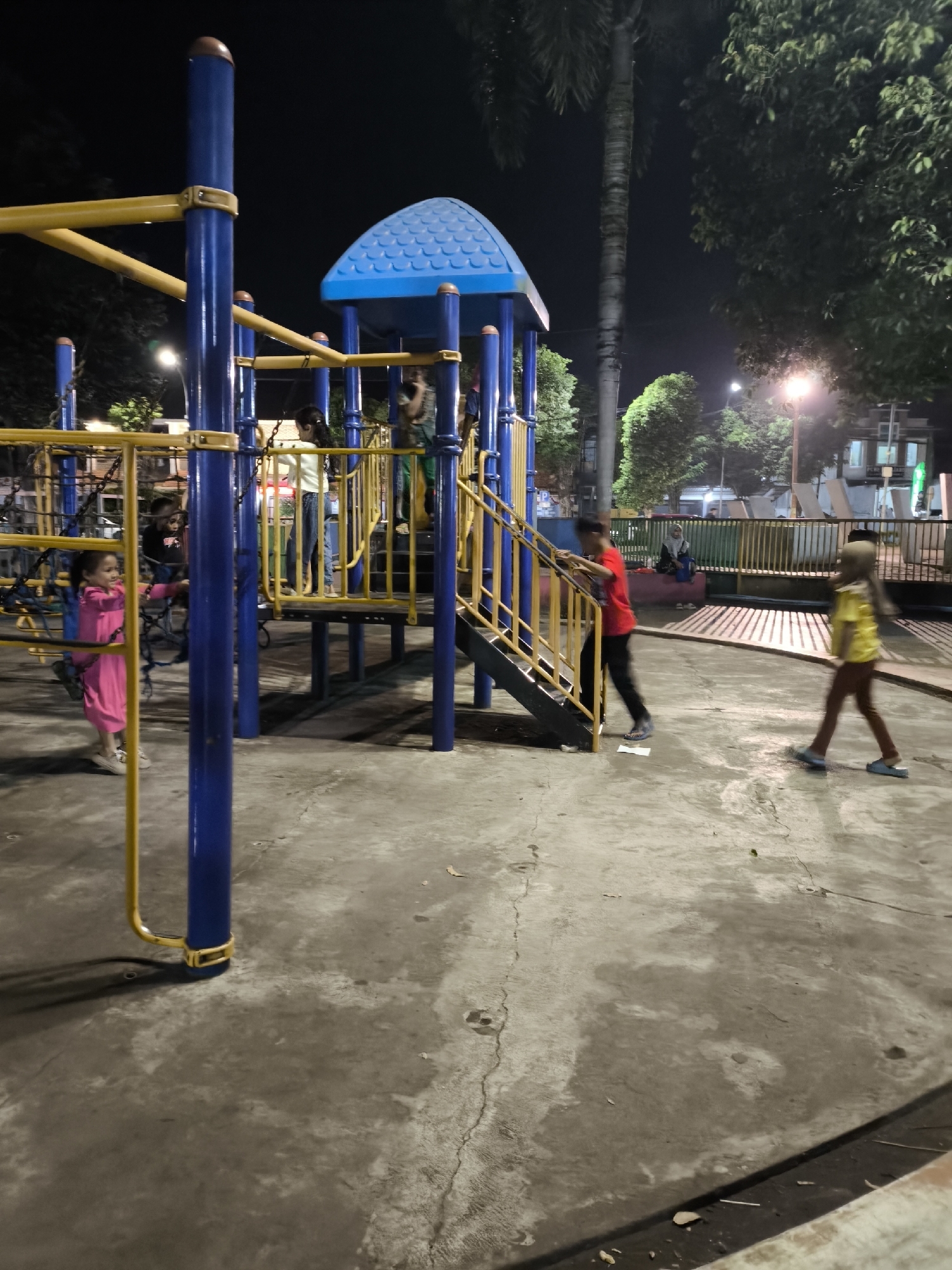 The image depicts a playground at night in Lumajang, Indonesia, illuminated by artificial lights. A blue and yellow metal playground structure with a blue roof is the central focus. Several children are present. On the left, a young child in a pink dress is on a swing. In the center, a child in a white shirt and blue jeans stands on the playground, and another child in a black shirt is visible behind them. To the right, a child in a red shirt is climbing a staircase of the playground, while a child in a yellow shirt walks away from the structure. The ground is paved with concrete. In the background, there are trees and buildings, with some streetlights visible.