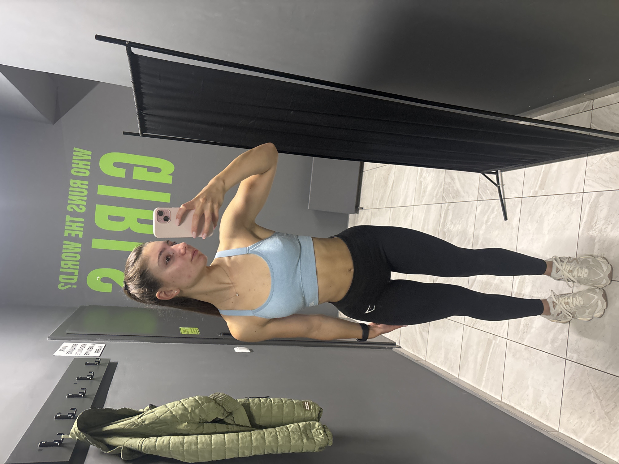 A young woman with brown hair pulled back in a ponytail is taking a selfie in what appears to be a locker room or changing area. She is wearing a light blue sports bra and black leggings, with white sneakers. To her left, a green jacket hangs on a row of hooks mounted on a grey wall. A large black screen stands to her right. In the background, bright green lettering on the grey wall reads, "WHO RUNS THE WORLD? GIRLS". The floor is tiled with a light grey and white marble pattern. The setting appears to be a gym or fitness facility. The image was taken in Varna, Bulgaria.