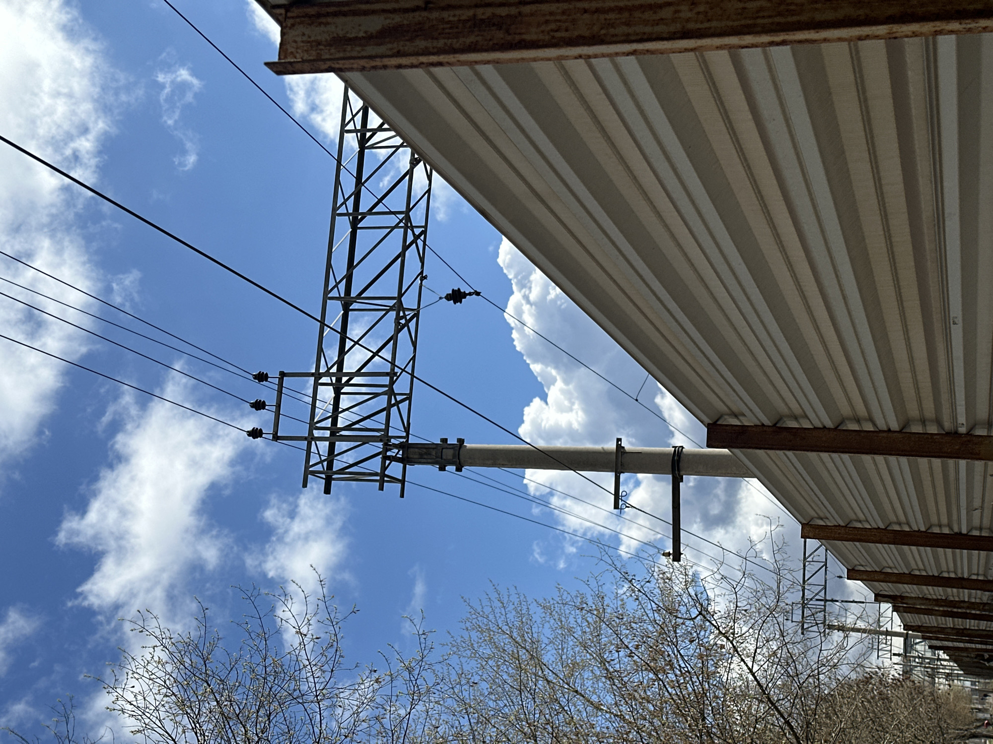 Looking up from below, the image shows an overhead power line structure in Moscow, Russia. The sky is bright blue with scattered white clouds. Bare trees with thin branches are visible in the foreground, suggesting early spring. The power lines are strung between a steel tower and a concrete pole which extends through a metal awning above the viewer's position. The awning is made of corrugated metal sheets, with visible rusted support beams. The perspective is angled, making the tower and wires appear to be converging upwards.