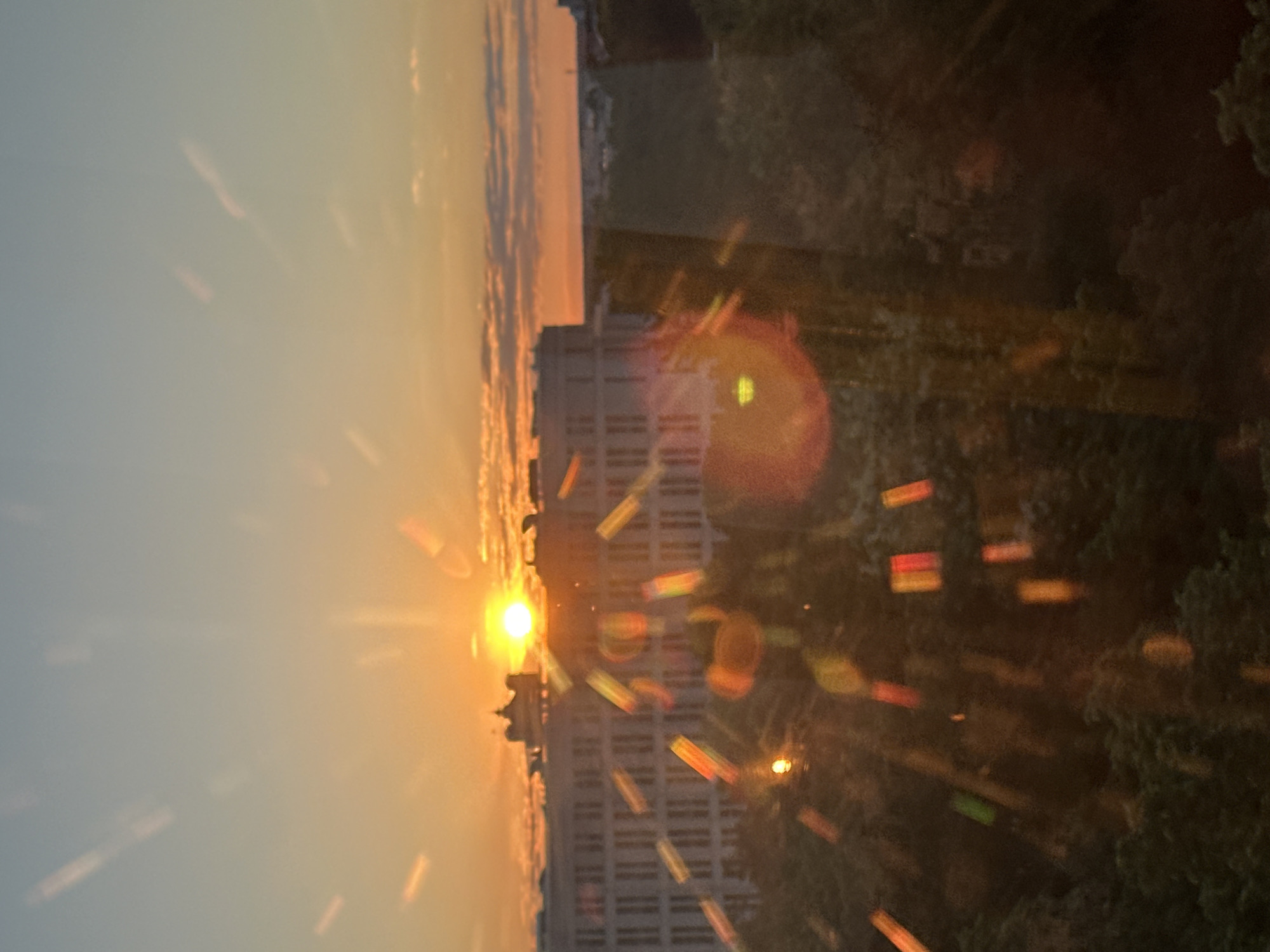 This image depicts a sunrise or sunset over a cityscape in Minsk, Belarus, viewed from an elevated position. The sky presents a warm gradient, transitioning from a soft blue-grey at the top to vibrant orange and yellow hues near the horizon, indicating the low position of the sun. The sun itself, a brilliant yellow orb, is situated towards the left of the frame, emitting strong radial lens flares. A prominent circular orange-red flare is visible on the right side of the image, and numerous scattered rectangular, multi-colored light artifacts suggest reflections or internal lens effects, possibly from viewing through a window.
Silhouetted against the bright sky are the dark outlines of multi-story buildings in the middle ground, with one structure showing distinct rectangular windows. Dense, dark green tree canopies are visible on the right and extending towards the bottom of the frame, providing a stark contrast to the illuminated sky. The time of day is clearly either early morning or late evening. The weather appears clear with some atmospheric haze. There is no visible text in the image. The scene conveys a sense of calm and natural beauty.