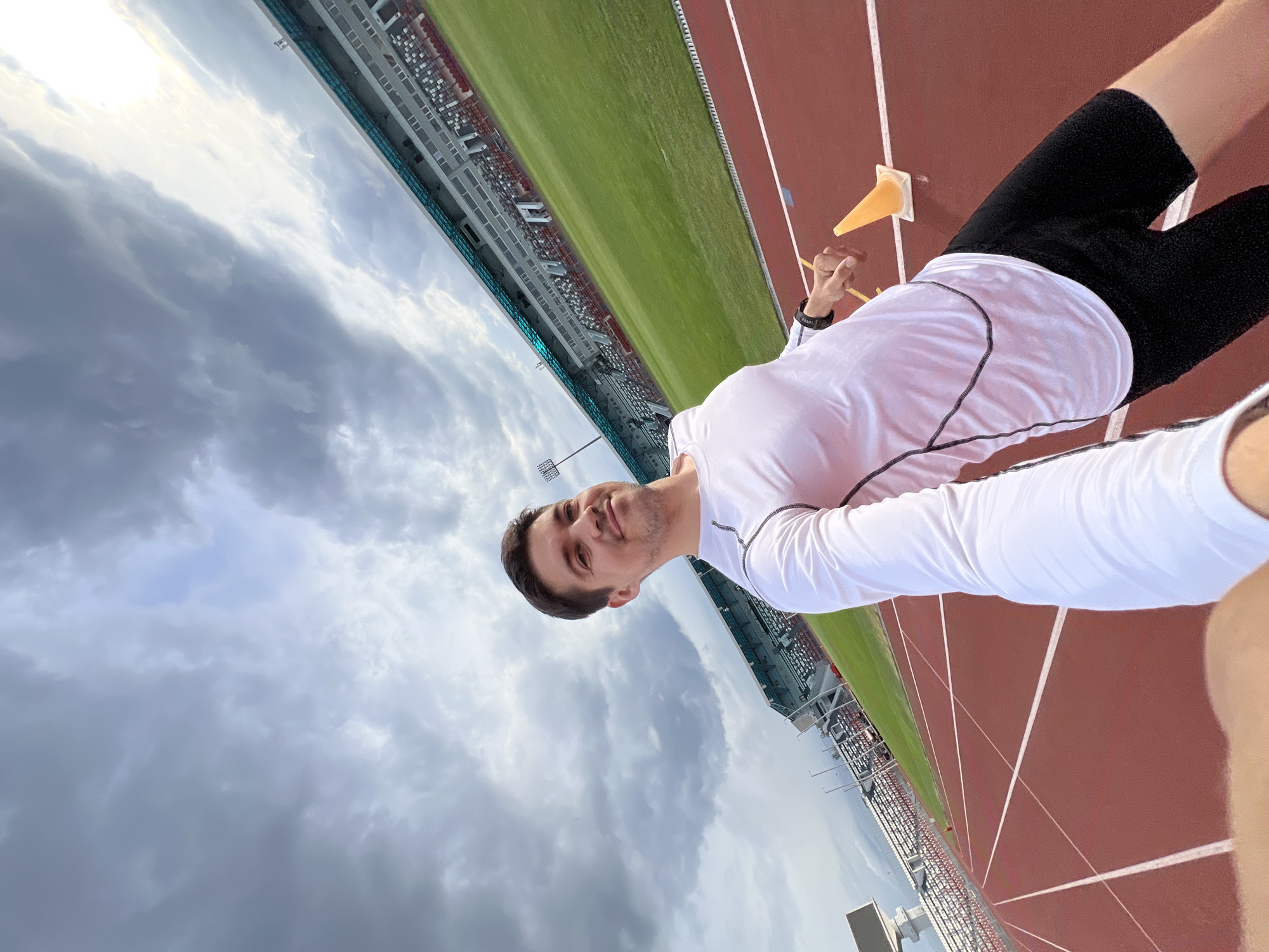 A medium eye-level outdoor shot, rotated slightly to the right, depicts a man on a running track. He is wearing a white long-sleeved shirt with black trim and black athletic shorts. A small orange cone rests on the track behind him. The track is composed of dark red lanes separated by white lines, and it runs adjacent to a green field. In the background, stadium seating rises in tiers, suggesting an athletic venue. The sky is overcast with heavy gray clouds. Based on the design of the stadium, it is likely located in Kazan, Russia. The man is smiling and facing the camera, which he may be holding at arm's length in a selfie pose.