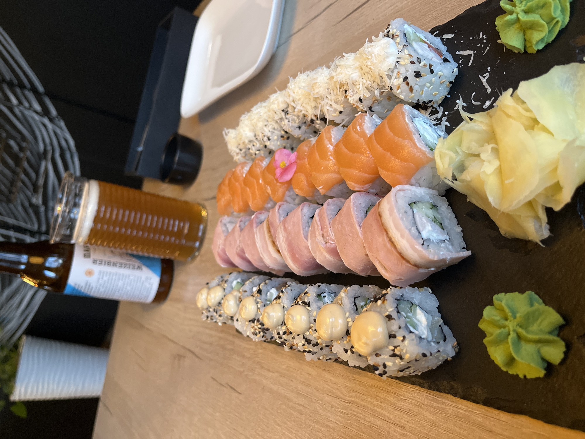 A top-down view captures a generously portioned platter of various sushi rolls and accompanying beverages, arranged on a wooden table. The central focus is a long black rectangular serving board holding several rows of maki sushi.
The sushi varieties include:
- Rolls topped with slices of vibrant orange salmon sashimi. One of these rolls is adorned with a small pink flower.
- Rolls covered in a fine, white, shredded topping, possibly tempura flakes or cheese.
- Rolls wrapped in thin, light pink/reddish slices of fish.
- Rolls sprinkled with black and white sesame seeds, each featuring dollops of a creamy, light yellow sauce on top.
Accompanying the sushi on the black platter are two common condiments: a dollop of bright green wasabi paste and a pile of pale yellow pickled ginger.
To the left of the platter, a tall glass filled with amber-colored beer, featuring a foamy head, is visible. Below the glass, a brown glass bottle is partially seen, with a label that clearly displays the word "WEISENBIER".
In the background, a white ceramic dish or bowl and a small black cup or bowl are present, resting on the wooden table. The immediate background is dark, suggesting indoor dining at a restaurant or home in Izhevsk, Russia. The overall scene depicts a delightful meal setting.