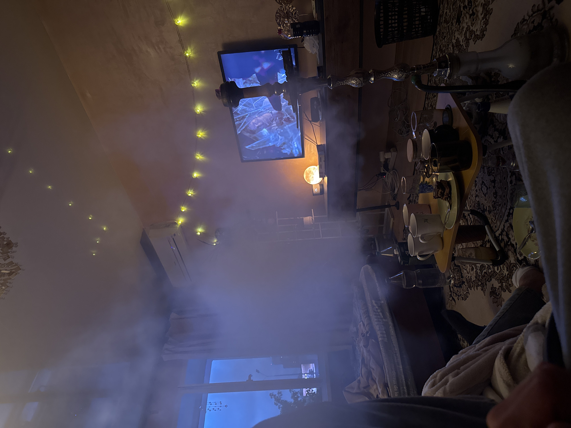 The image depicts an indoor room, likely a residential living space or bedroom, filled with dense white smoke or vapor, particularly prominent in the midground and left side. In the lower-left foreground, a person is partially visible, seated on the floor with their lower torso, legs, and left hand in view. They wear grey trousers and white socks on their visible foot, with a light-colored blanket draped over their lap. They appear to be facing towards the right side of the room. To the left, a large window reveals a dark blue twilight or nighttime exterior. Distant streetlights and what appears to be a building or vehicle can be faintly seen through the haze. A decorative string of clear beads hangs in the upper-left pane. White curtains frame the window, and beneath it, a bed with a visible mattress (featuring upside-down text "Bonheur") and disheveled light-colored bedding is present. The right side of the room features a light brown or beige textured wall. A flat-screen television is mounted on this wall, displaying a blue-toned image of a person's face, possibly from a movie or television show. Below the TV is a dark wooden console or desk, on which a spherical, glowing orange 'moon' lamp sits beside a small digital clock displaying "26." Cables are visible beneath the console. Above the TV and extending across the ceiling, numerous small yellow LED fairy lights illuminate the space. An air conditioning unit is visible on the wall to the left of the TV. In the central foreground, a small, yellow, rectangular lap table is positioned on a dark, intricately patterned rug. On the table are several items: a white ceramic mug, a black ceramic mug, a beige ceramic mug, an empty clear glass, a lidded glass jar, and a small plate holding dark brown pastries (possibly brownies). A large clear plastic water bottle and a small white carton are on the floor adjacent to the table. Prominently placed in the midground, between the viewer and the TV, is a tall, ornate silver hookah with dark red or purple accents on its bowl. Its clear glass base is visible, and it is actively producing the smoke that fills the room. To the far right, a black, open-weave plastic trash can is visible on the rug. The floor is covered by a dark, ornate rug with floral or paisley patterns. The overall lighting is dim,