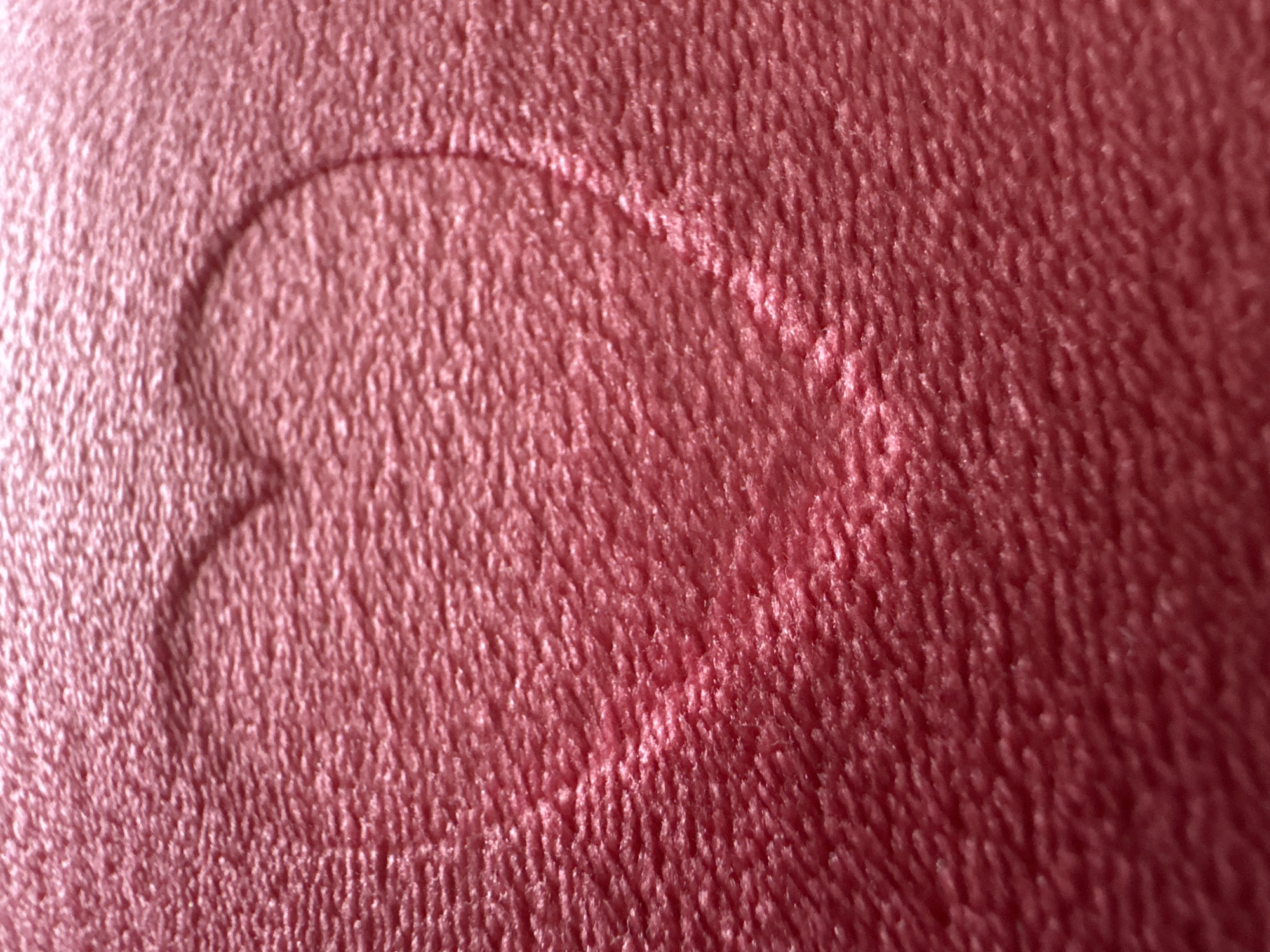 The image presents a close-up, high-angle view of a textured fabric surface, predominantly colored in a warm reddish-pink or maroon hue. The material appears soft and plush, possibly fleece or velvet, exhibiting a dense, finely corrugated texture of short fibers.
A distinct heart shape is embossed or pressed into the fabric, positioned towards the center-left of the frame. This impression creates an area that appears lighter in shade and smoother or more compressed than the surrounding textured material, clearly outlining the heart. The lighting is soft and diffused, highlighting the contours of the heart and the minute details of the fabric's nap, creating subtle variations in light and shadow that emphasize its three-dimensional quality.
No people, other objects, or background elements are visible, making the fabric and the heart impression the sole focus. There is no discernible activity taking place, and the scene is static. No text is visible within the image. While the location context is Tallinn, Estonia, there are no visual cues in the close-up to indicate this specific location.