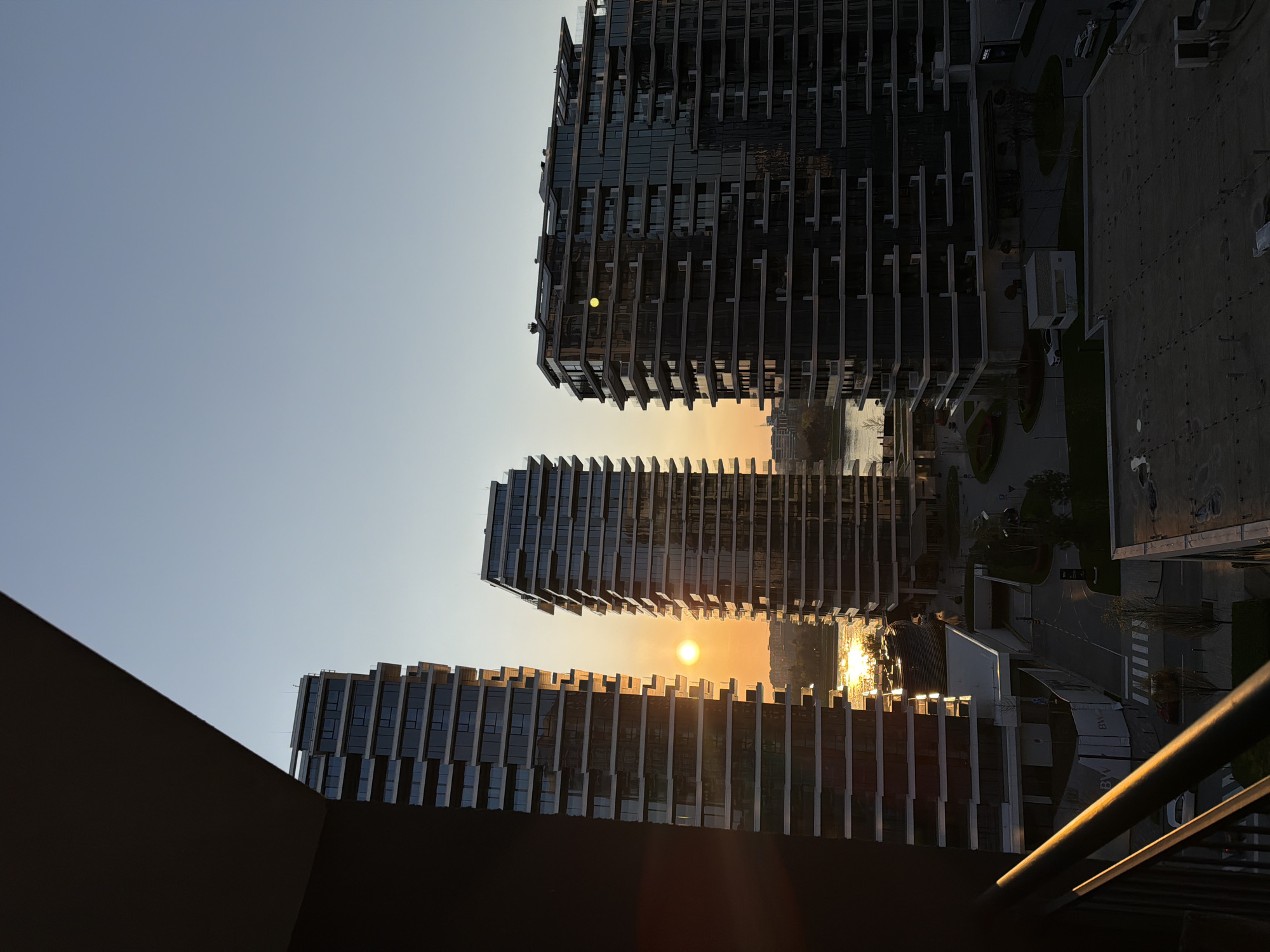 The image shows a view of buildings in Belgrade, Serbia, at sunset. The sun is positioned between two buildings, creating a glowing effect. There are three prominent buildings with a modern architectural style. The building in the foreground has horizontal lines on its facade. A railing is visible on the right side of the frame, suggesting the photo was taken from a balcony or elevated position. The sky is light blue, fading towards a yellow-orange horizon where the sun is setting. A body of water is visible behind the buildings, reflecting the sunlight.