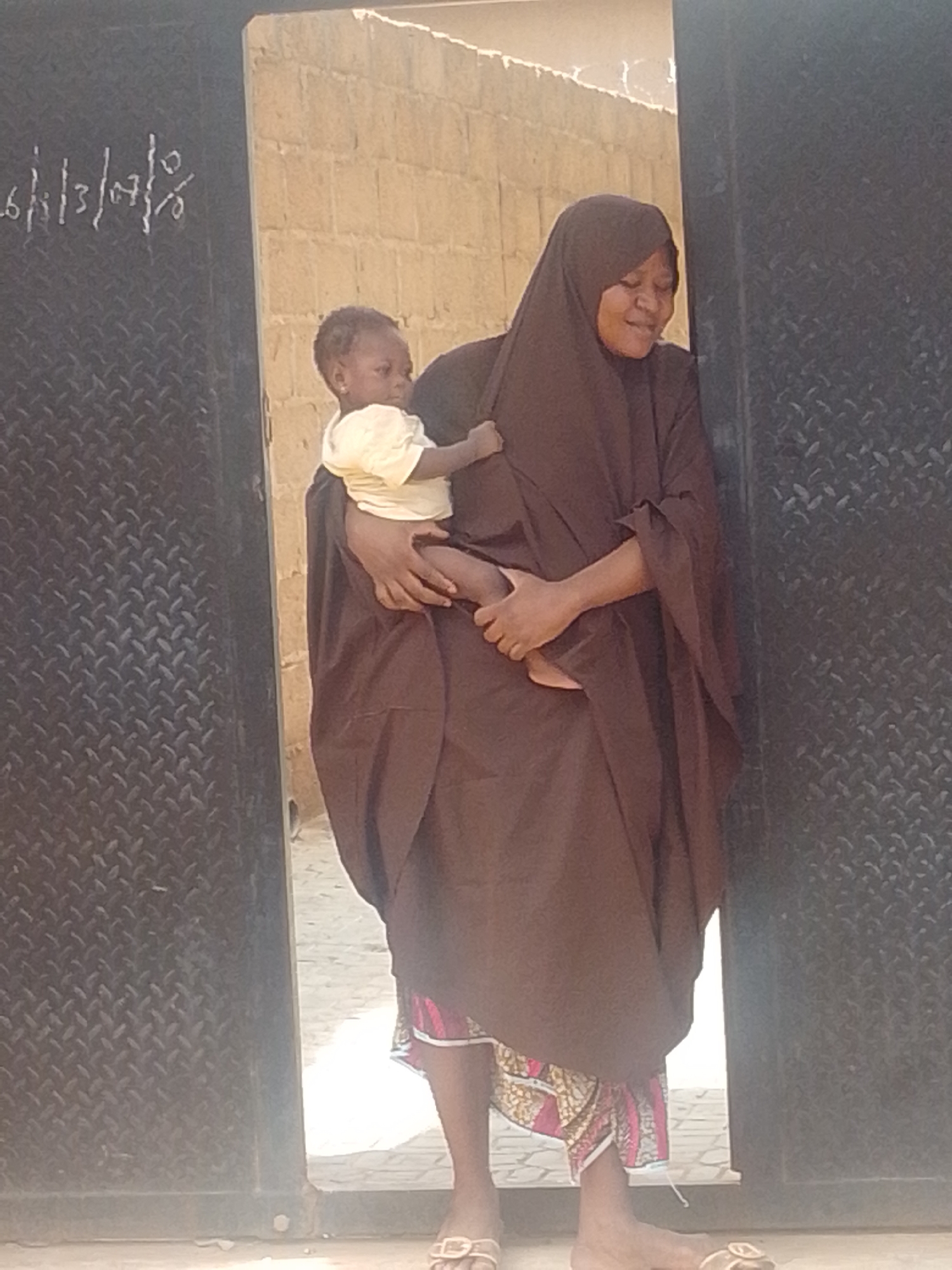 A woman wearing a brown hijab and dress holds a baby wrapped in a brown cloth. The baby wears a light yellow shirt. The woman is smiling and looking down at the baby. They are standing in front of a metal gate with a diamond plate pattern. A brick wall is visible in the background. There is writing on the gate that appears to be a date: "16/13/07/8".