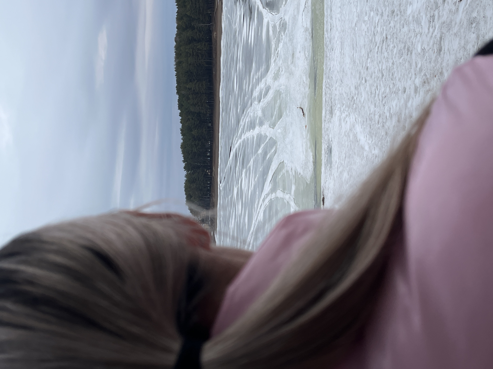The image shows a person with long, blonde hair and a pink garment standing near a body of water. The water is partially covered in ice, with a snow-covered shoreline. In the background, a dense forest is visible against a cloudy sky. The location is Unknown, Unknown.