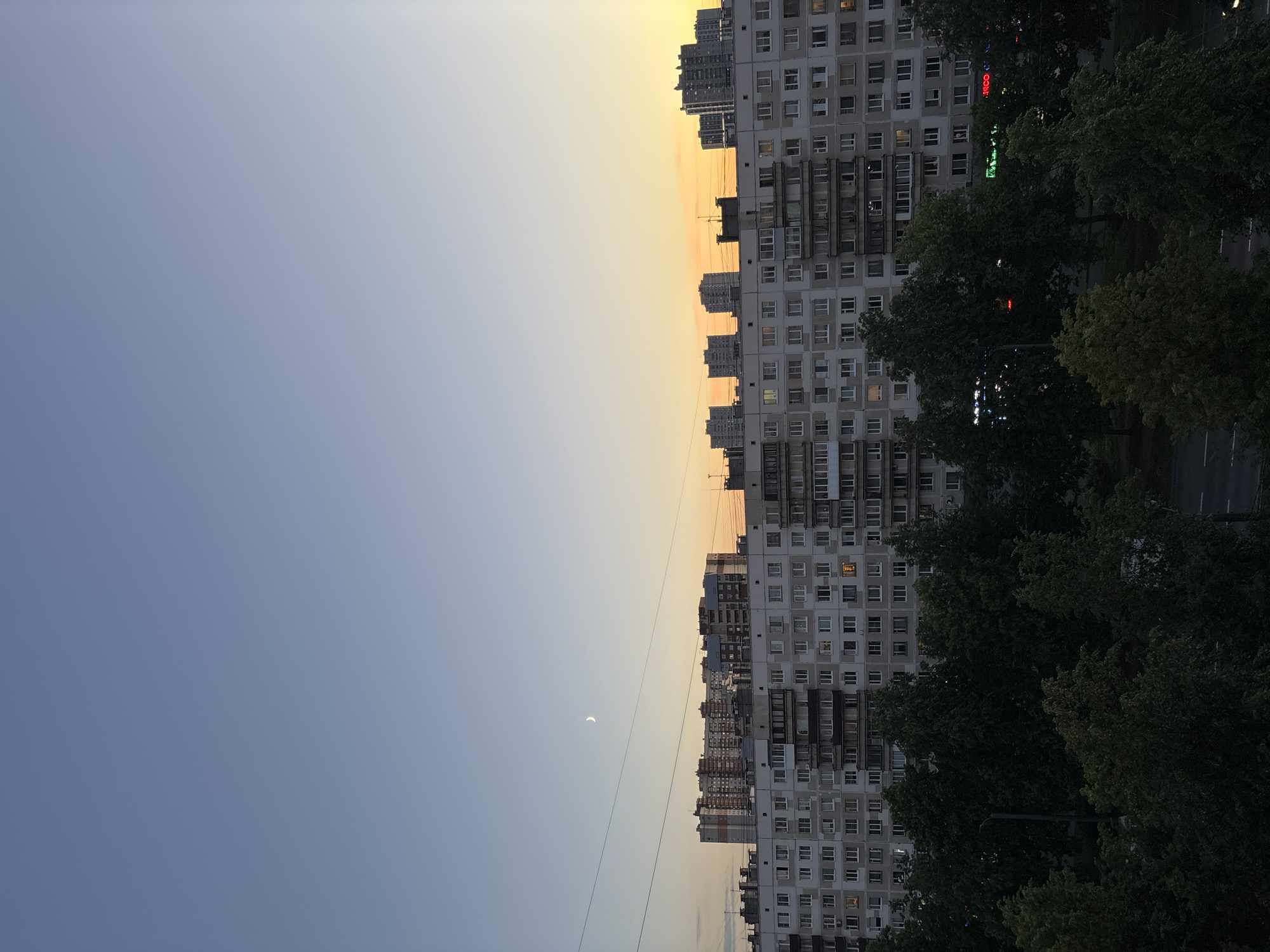 The image shows a twilight view of a residential area in an urban setting. In the foreground, dense foliage of trees obscures the lower portion of the scene, with hints of a street and possibly a car visible. Behind the trees, a multi-story apartment building with a pale facade and numerous windows dominates the mid-ground. Some windows are illuminated, suggesting occupancy. Above the apartment building, several other taller buildings are visible, receding into the distance. The sky transitions from a clear, pale blue at the top to a warm, orange-yellow hue near the horizon, indicating either sunset or sunrise. A thin crescent moon is visible in the upper left portion of the sky. Electrical wires are strung across the sky in the upper mid-ground. The overall impression is of a quiet evening in a densely populated urban environment.
