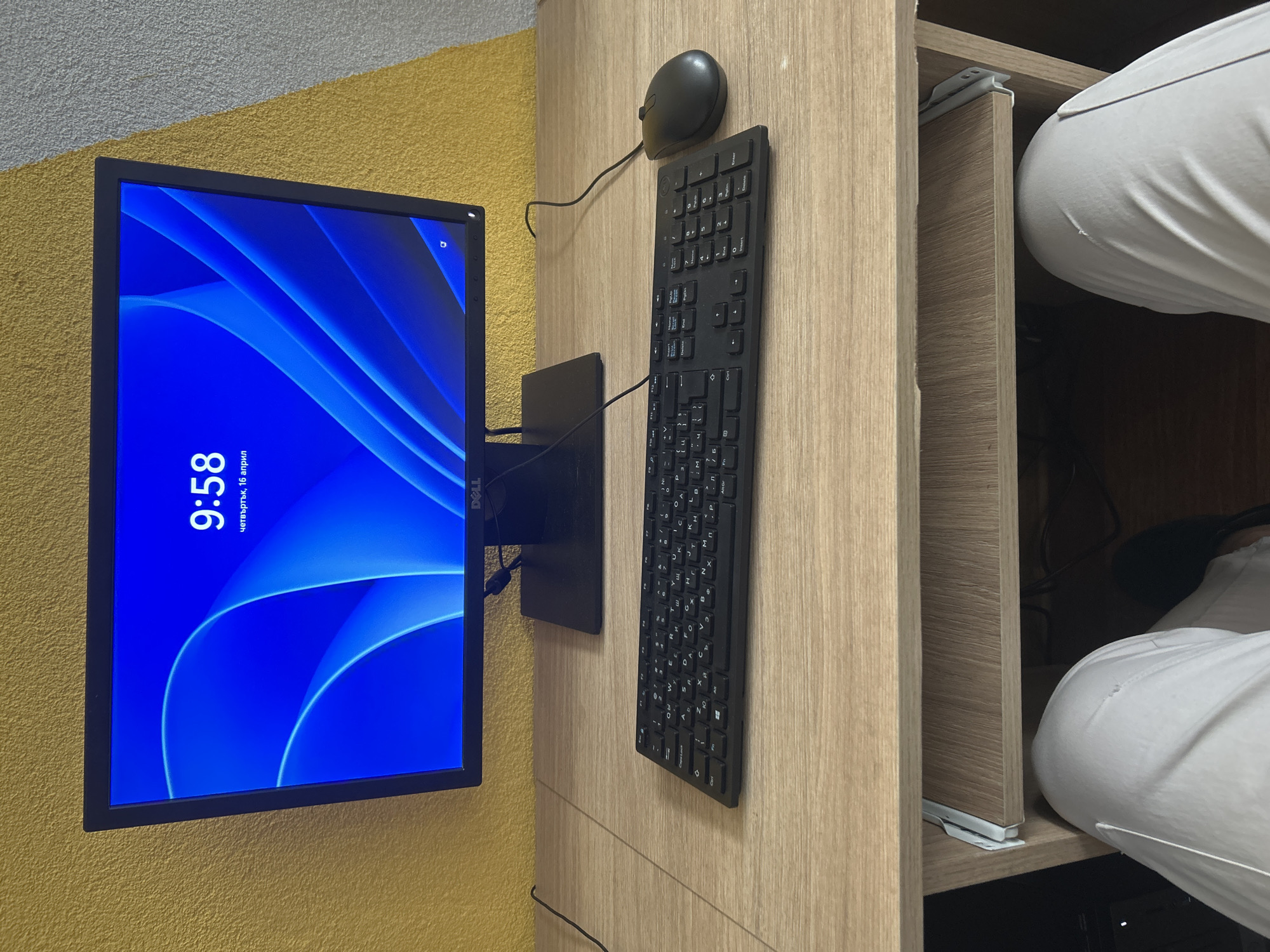 The image shows a computer setup in an indoor setting. A black Dell monitor displays a blue abstract wallpaper with the time "9:58" and the date "четвъртък, 16 април." The monitor is mounted on a yellow wall. To the right of the monitor, on a light wood-grain surface, are a black computer mouse and a black Dell keyboard. The right side of the frame includes a partially open light wood-grain drawer with a person wearing white clothing visible inside. Although the date displayed on the screen is April 16th, the year is unknown. The location is assumed to be Zlatograd, Bulgaria, but the image does not provide concrete evidence.