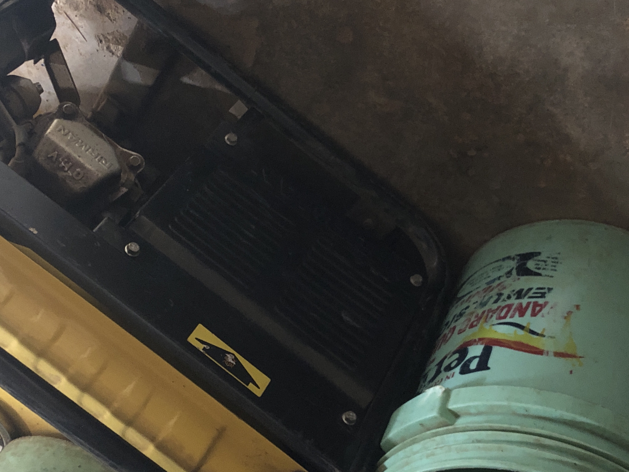 The image shows a portion of a piece of machinery, potentially a compactor, with a yellow fuel tank and a black engine cover. The engine cover has a warning sticker with a black pictogram on a yellow background. Next to the machinery is a light green bucket with markings. The background is a mottled brown color, possibly concrete or dirt. The exact location is unknown.