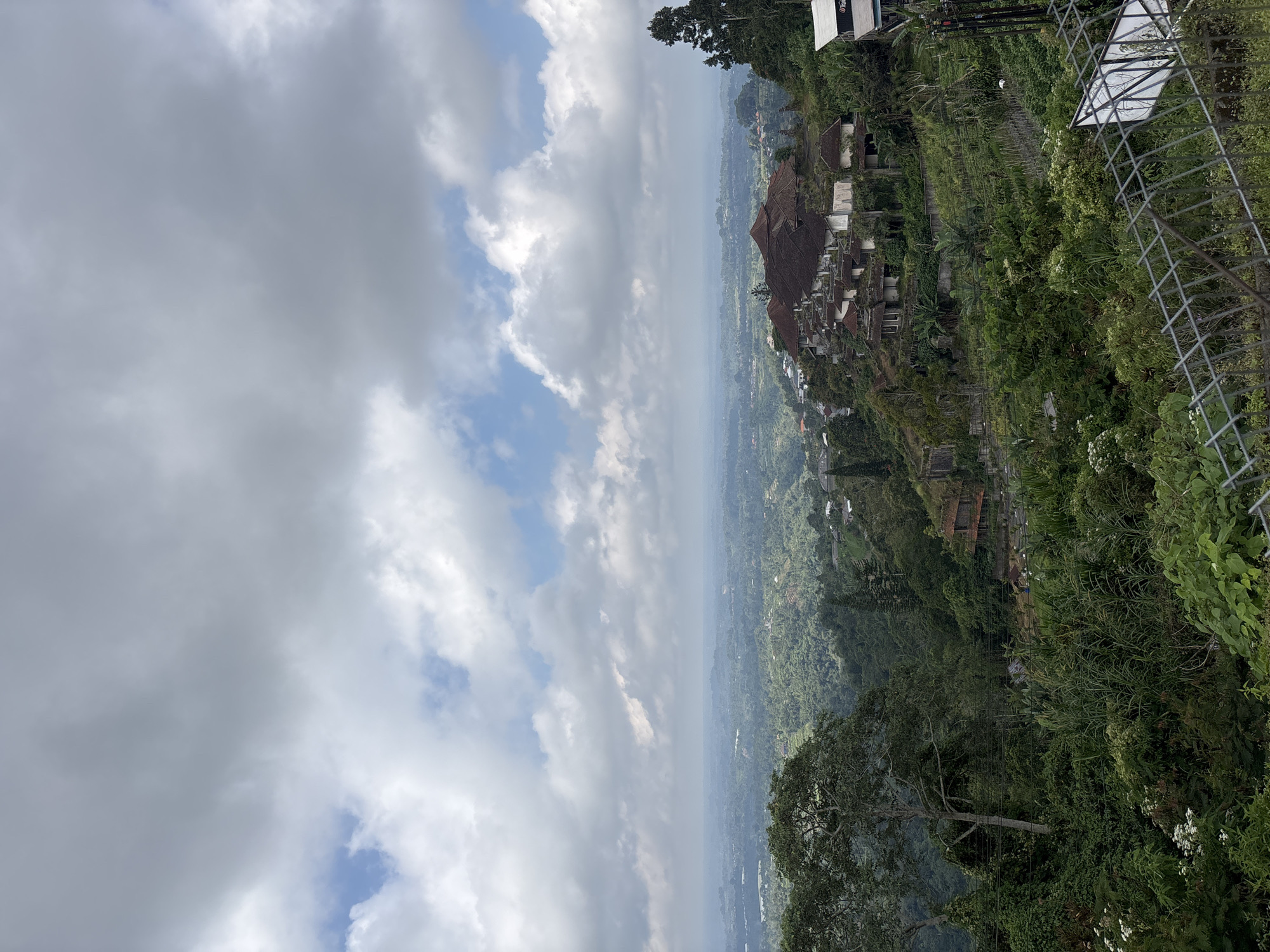 The photograph depicts a landscape in Baturiti, Indonesia, taken from an elevated perspective. The foreground consists of dense green vegetation, including trees and foliage. Beyond this, a panoramic view reveals the town’s layout, showcasing buildings with traditional architecture and terraced structures embedded within the terrain. In the distance, a vast expanse of land extends towards the horizon, fading slightly due to atmospheric haze. The sky occupies a significant portion of the image, dominated by a mix of white and gray clouds. A metal fence is visible on the right side of the image, adding a man-made structure to the natural scenery.