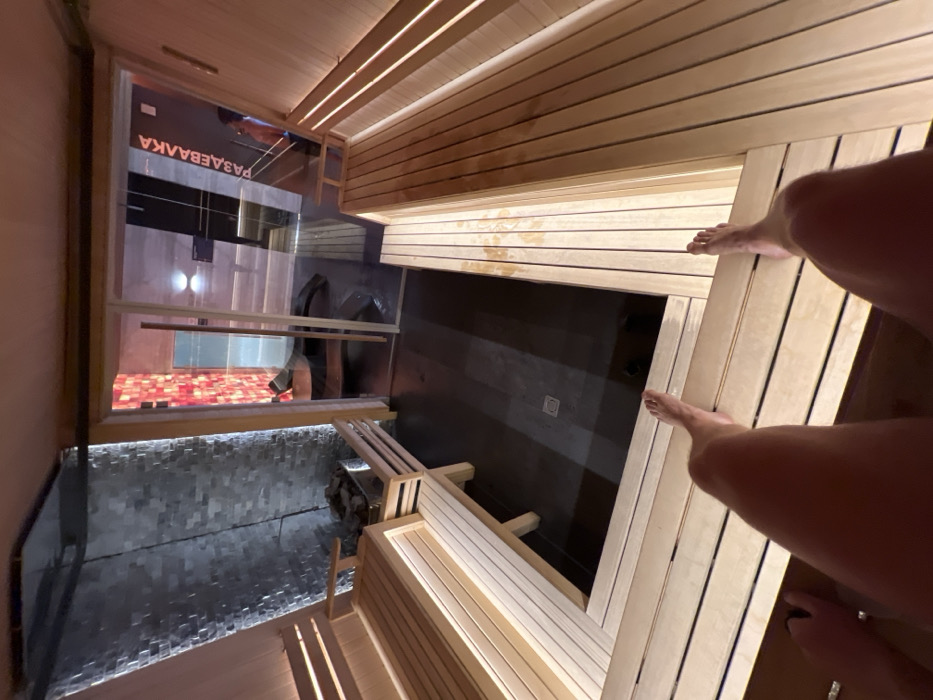 The image captures an interior view of what appears to be a modern sauna or spa facility, characterized by wooden paneling and dim, atmospheric lighting. The perspective is from a high angle, looking downwards, likely from an upper bench. In the immediate foreground, a person's bare legs and feet are visible, resting on wooden slats, indicating they are seated or reclining within the sauna.
The space features a warm wooden aesthetic with multiple tiers of benches. A prominent glass wall separates the sauna area from an adjacent room. Through this glass, reflections are visible, and another person can be faintly seen in the background. The floor beyond the glass wall appears to be a dark, possibly tiled or concrete surface, leading to an area with reddish-toned floor tiles or lighting, and some dark objects resembling bags or towels.
To the left, a textured wall, possibly made of dark stone or mosaic tiles, is illuminated from below by integrated strip lighting, creating a soft glow. Adjacent to this wall, what looks like a sauna heater with stones is subtly visible. The main floor of the sauna, visible below the benches, is a dark, sleek material, possibly concrete or large tiles, with a drain present.
Visible text, reflected and inverted through the glass wall, reads "РАЗДЕВАЛКА," which translates from Russian to "CHANGING ROOM" or "CLOAKROOM." The overall ambiance is one of relaxation and warmth, typical of a spa or bathhouse setting in Saint Petersburg, Russia. The lighting suggests an indoor setting, likely evening or artificial light dominating.