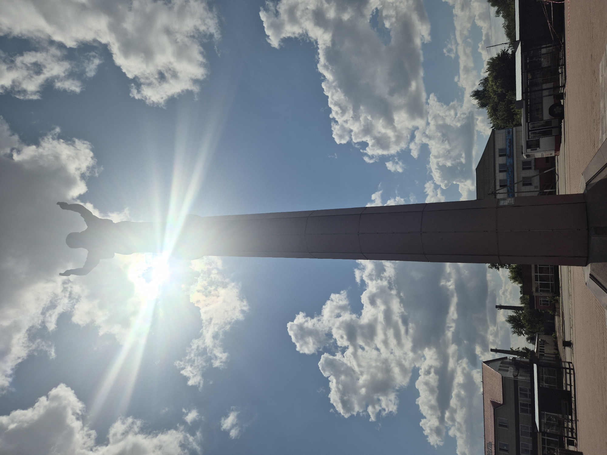 The image captures a statue of a person with outstretched arms, silhouetted against a bright, sunlit sky filled with fluffy white clouds. The statue stands atop a tall, cylindrical monument, casting a shadow. In the foreground, there is a paved area with red brickwork. To the left and in the background, there are buildings with red and green roofs, and a bus. The scene appears to be an outdoor public space, possibly a plaza or square, in the city of Kokshetau, Kazakhstan. The lighting suggests it is daytime, with the sun creating a halo effect around the statue.