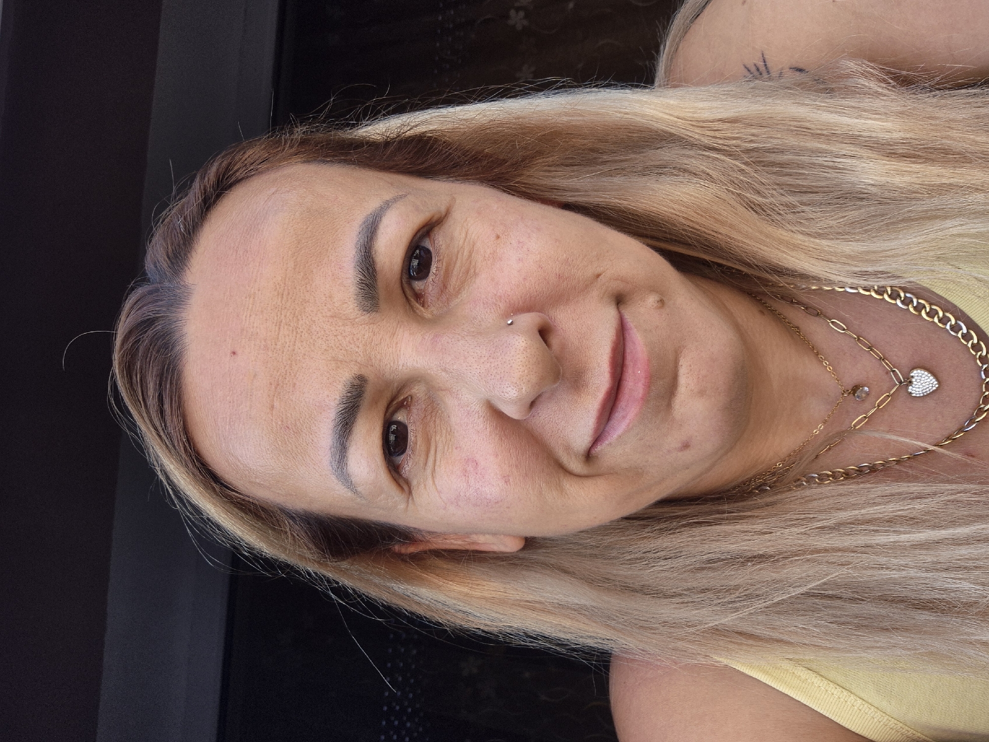 A medium close-up outdoor selfie shows a fair-skinned person with blonde hair, a nose ring, and several gold necklaces. They are looking at the camera with a slight smile. The background is dark and out of focus, suggesting it might be a window or doorway with some light filtering through. The person is wearing a yellow tank top.