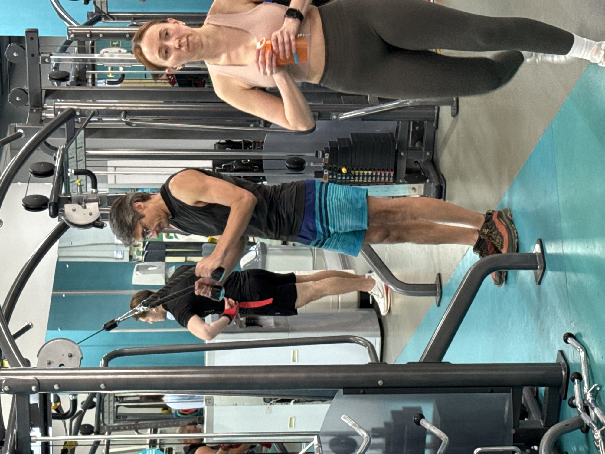 The image depicts an indoor gym scene, likely situated in Yekaterinburg, Russia, featuring three individuals engaged in or pausing from physical activity during what appears to be daytime.
On the left side of the frame, a man with short, dark hair and glasses, wearing a dark gray tank top and blue and black striped athletic shorts, is actively performing an exercise on a cable machine. He grips a handle with both hands, positioned for a pulling or pushing movement. He is wearing athletic shoes with brown and orange detailing.
Behind him, a large mirror reflects another man who is also exercising. This reflected individual is wearing a black short-sleeved top and black shorts with a red accent, and appears to be using gym equipment.
On the right side of the frame, a woman with light brown hair stands, looking directly at the camera with a neutral expression. She is dressed in a light pink sports bra, dark gray leggings, and white athletic socks. She holds an orange and clear water bottle at her waist. A black smartwatch is visible on her left wrist.
The gym environment includes various pieces of dark-colored exercise equipment, such as multiple cable machines with visible weight stacks. The floor is a combination of light gray and a bright blue section. No specific text is clearly visible on any items of clothing or equipment.