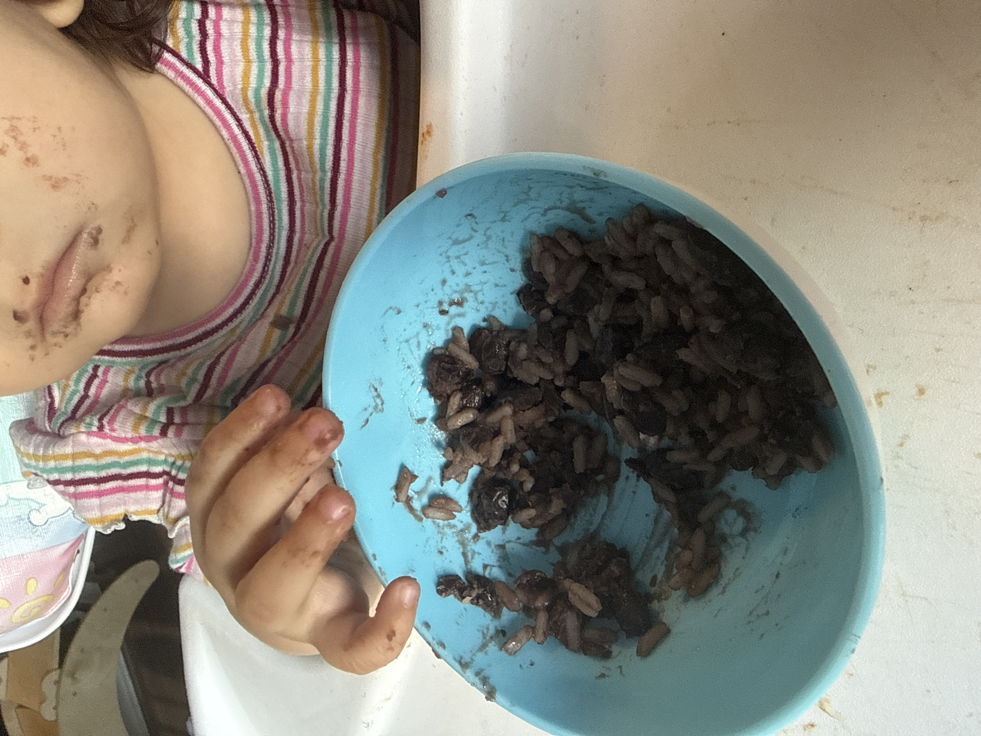 A child sits in a high chair, facing left with a blue bowl of dark grains, possibly rice, in front of them. The child's face and hand are smudged with the food. The child wears a multi-colored striped shirt. The high chair is white and partially visible.