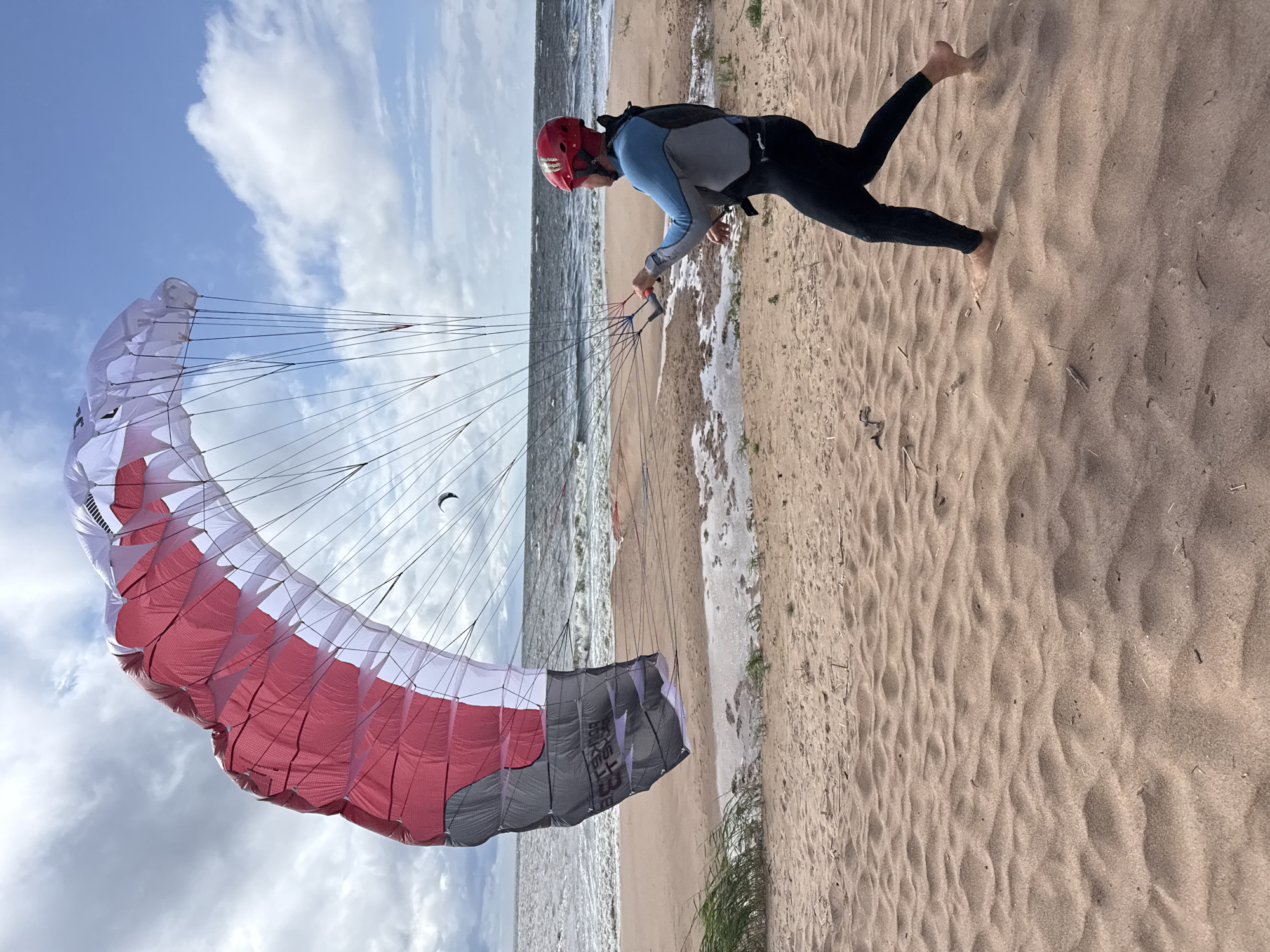A person in a red helmet and a blue and black wetsuit is on a sandy beach, preparing to kitesurf. They are holding onto the control bar of a large, red and white kite. The kite is inflated and angled upwards against a cloudy sky. The beach is light brown sand with some dry grass and debris. In the background, the sea has moderate waves, and a few other small kites are visible on the horizon. The scene appears to be the coast near Zelenaya Roshcha, Russia.
