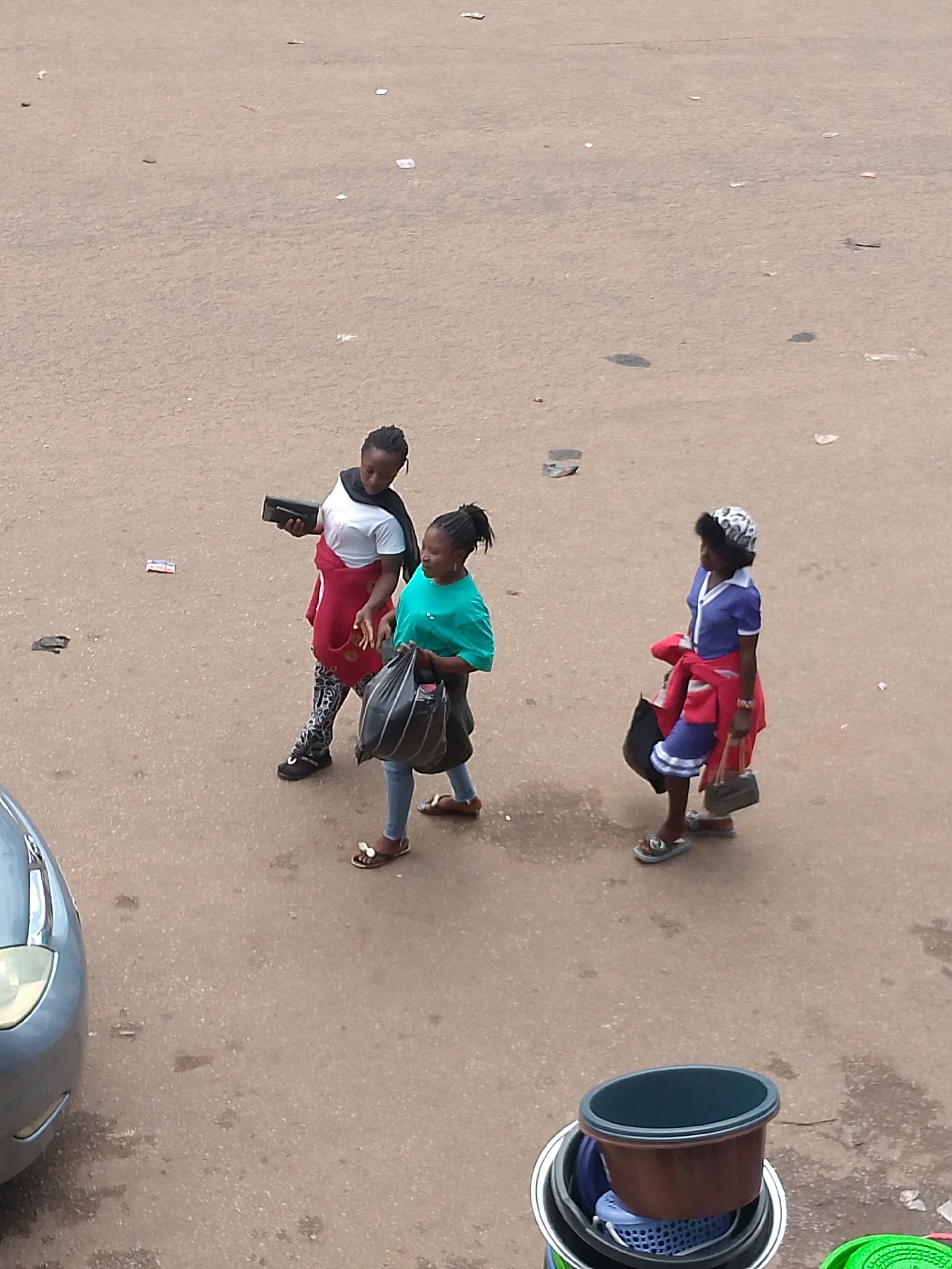 In Bukuru, Nigeria, three young women are walking on a paved street. The woman on the left wears a white shirt, a red jacket, and patterned leggings. She carries a black rectangular object in her left hand. The woman in the middle wears a teal shirt and blue jeans and carries a large black plastic bag in her right hand. The woman on the right wears a blue dress, a red jacket tied around her waist, and a patterned headscarf. She carries a black handbag in her left hand. In the foreground, several buckets are stacked on the ground. A car is partially visible on the left side of the image. The street is dirty and littered with small pieces of trash.