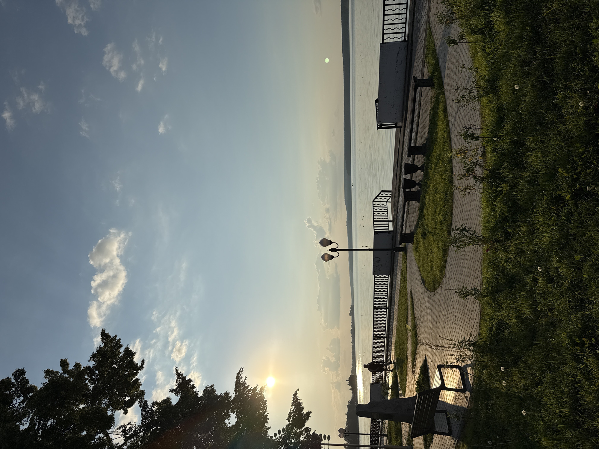 The image depicts a serene waterfront promenade in Votkinsk, Russia, during what appears to be a late afternoon or early evening. The sun, low in the sky, casts a warm golden glow, creating a gentle haze that softens the landscape. A lone figure, silhouetted against the bright light, walks along the paved pathway bordering a calm body of water, likely the Votka River or Lake Votkinsk.
The promenade is lined with a decorative black metal railing and interspersed with lampposts. Benches are placed along the path, some occupied by the shadows of the trees overhead. Lush green grass covers the area between the path and the water's edge, with a few dandelions dotting the foreground. Across the water, a distant treeline marks the horizon, and faint clouds drift across the pale blue sky. In the far distance, a few buildings are visible, suggesting the presence of the city. The overall atmosphere is one of tranquility and natural beauty.
