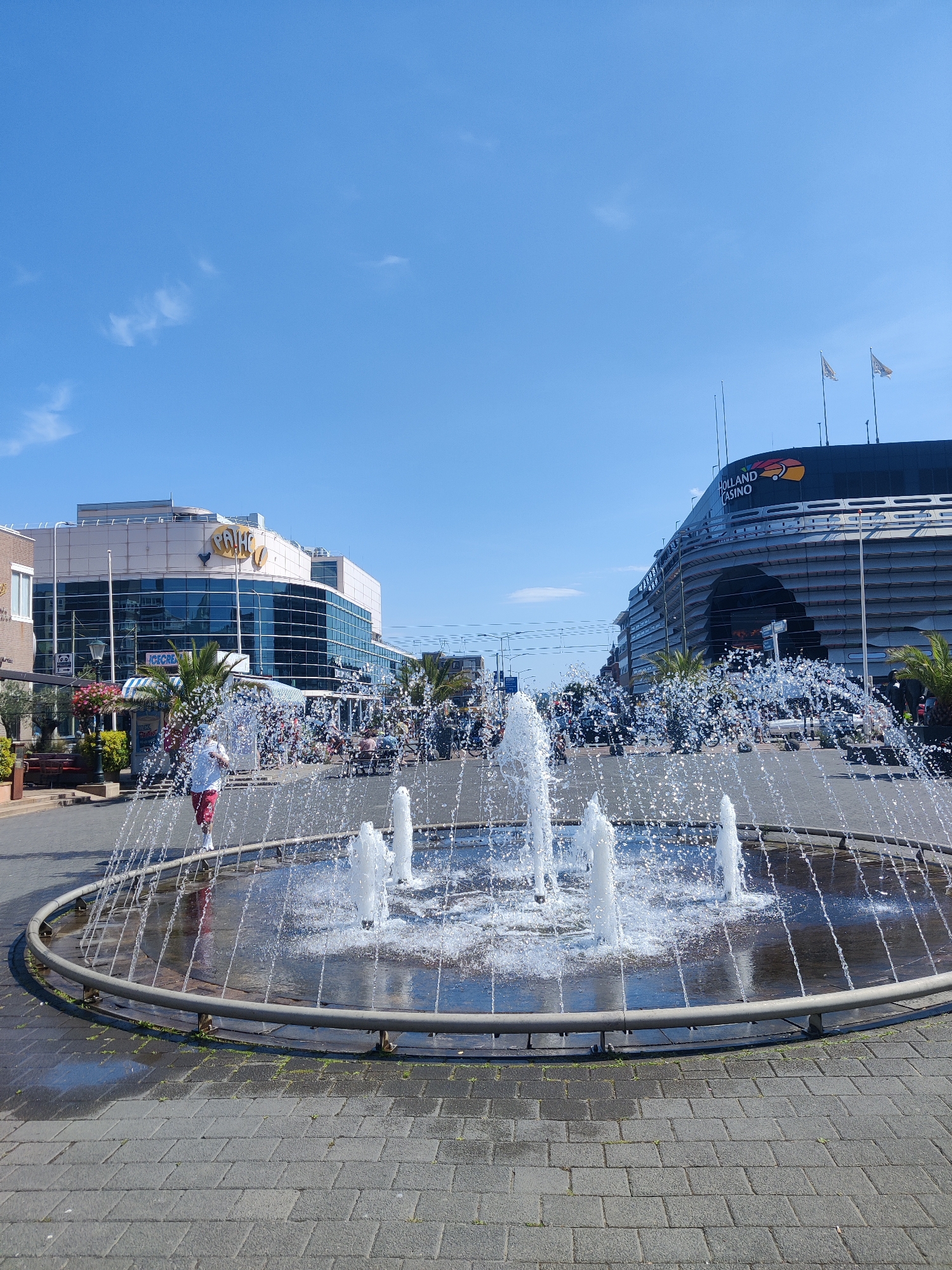 This is a street-level photograph taken in front of the Holland Casino in Scheveningen, a district of The Hague, Netherlands. The foreground features a circular fountain with water jets spraying upwards and outwards, creating a shimmering effect. A person wearing a white t-shirt and red and white patterned pants is partially visible in the middle ground, running near the fountain.
To the left of the fountain is a modern building with a glass facade, featuring the word "PALACE" prominently displayed. There are palm trees and flower arrangements in front of this building. Beyond this, more of the city's architecture is visible.
To the right of the fountain, the large, curved structure of the Holland Casino is dominant. It has a dark facade and a prominent logo with the words "HOLLAND CASINO" and an orange and red graphic. Two flags are visible on poles above the casino.
The background consists of a clear, bright blue sky with a few wispy white clouds. The ground in the foreground and mid-ground is paved with grey interlocking bricks. The overall impression is a sunny day in a lively urban plaza.