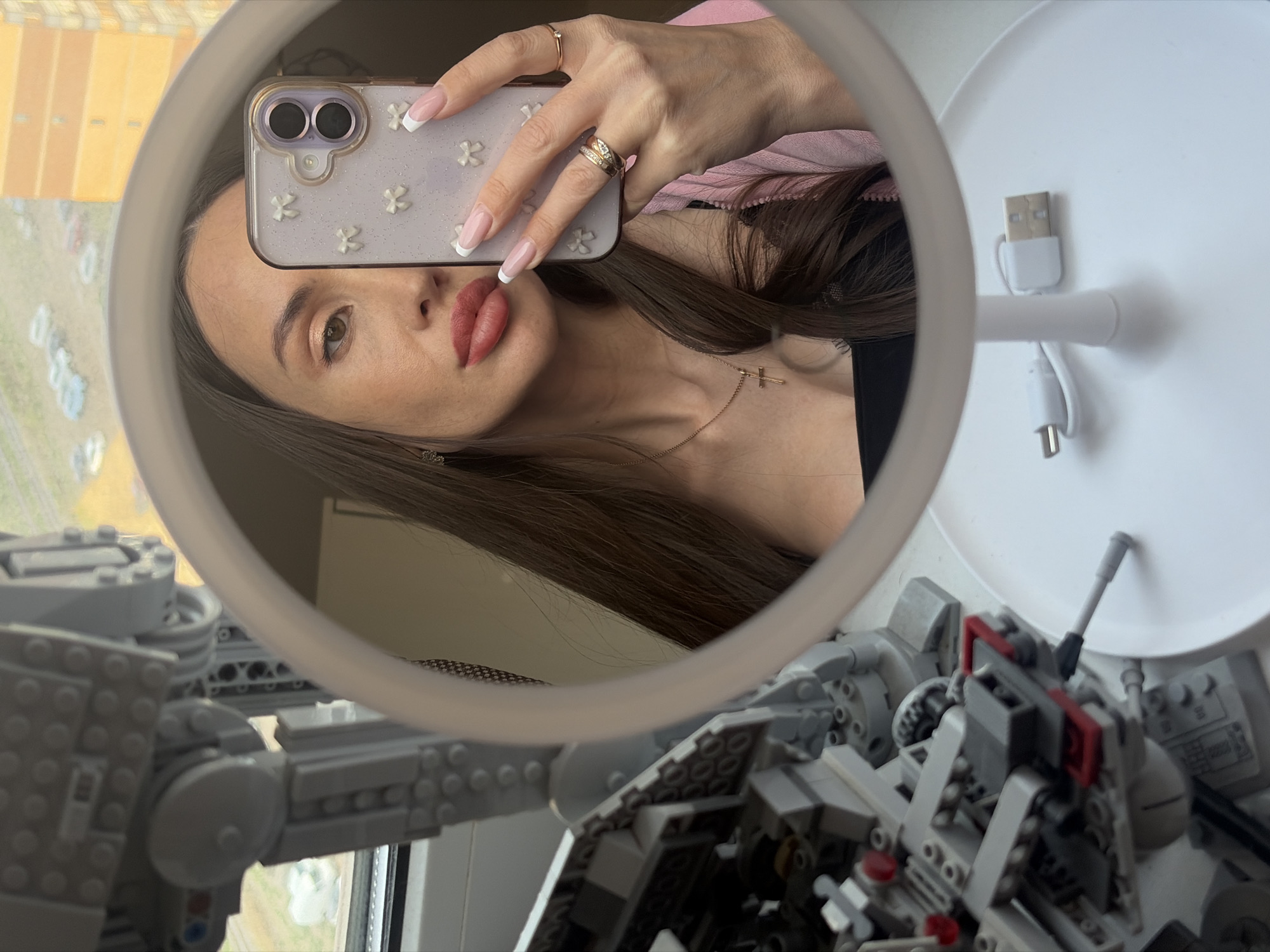 A woman is taking a selfie in a round mirror. The reflection shows her face, left eye visible, lips, and part of her hair. She is holding a phone with a light-colored case decorated with flower patterns. Her manicured fingers with rings are visible. She wears a pink jacket and a necklace with a cross pendant.
The mirror is on a white stand with a USB cable attached. Below the mirror, part of a Lego model of an AT-AT is visible. A blurred outdoor scene with yellow buildings is visible through a window behind the Lego model. The building suggests the location to be in Saint Petersburg, Russia.