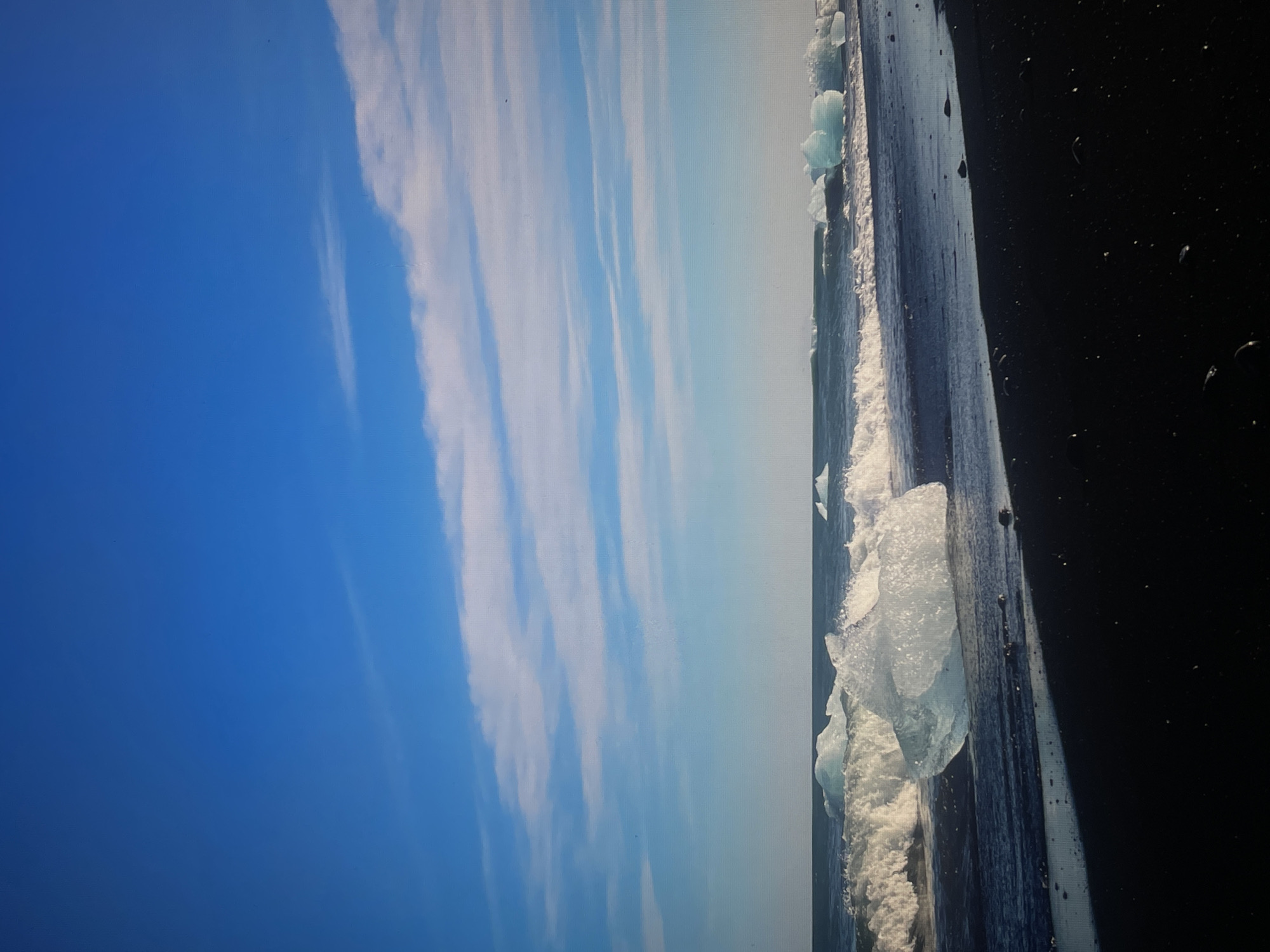 The image shows a scene of water, ice, and sky. On the right side of the image is what appears to be the hull of a dark-colored ship with ice formations and water splashing against it. The ice formations are translucent and irregularly shaped. To the left of the ship's hull, the scene transitions into a view of the sky, which is blue with streaks of white clouds. The sky is brighter closer to the horizon. The image is oriented in a manner that the sky occupies a larger portion of the left side, with the ship hull dominating the right. The location is Unknown, Unknown.
