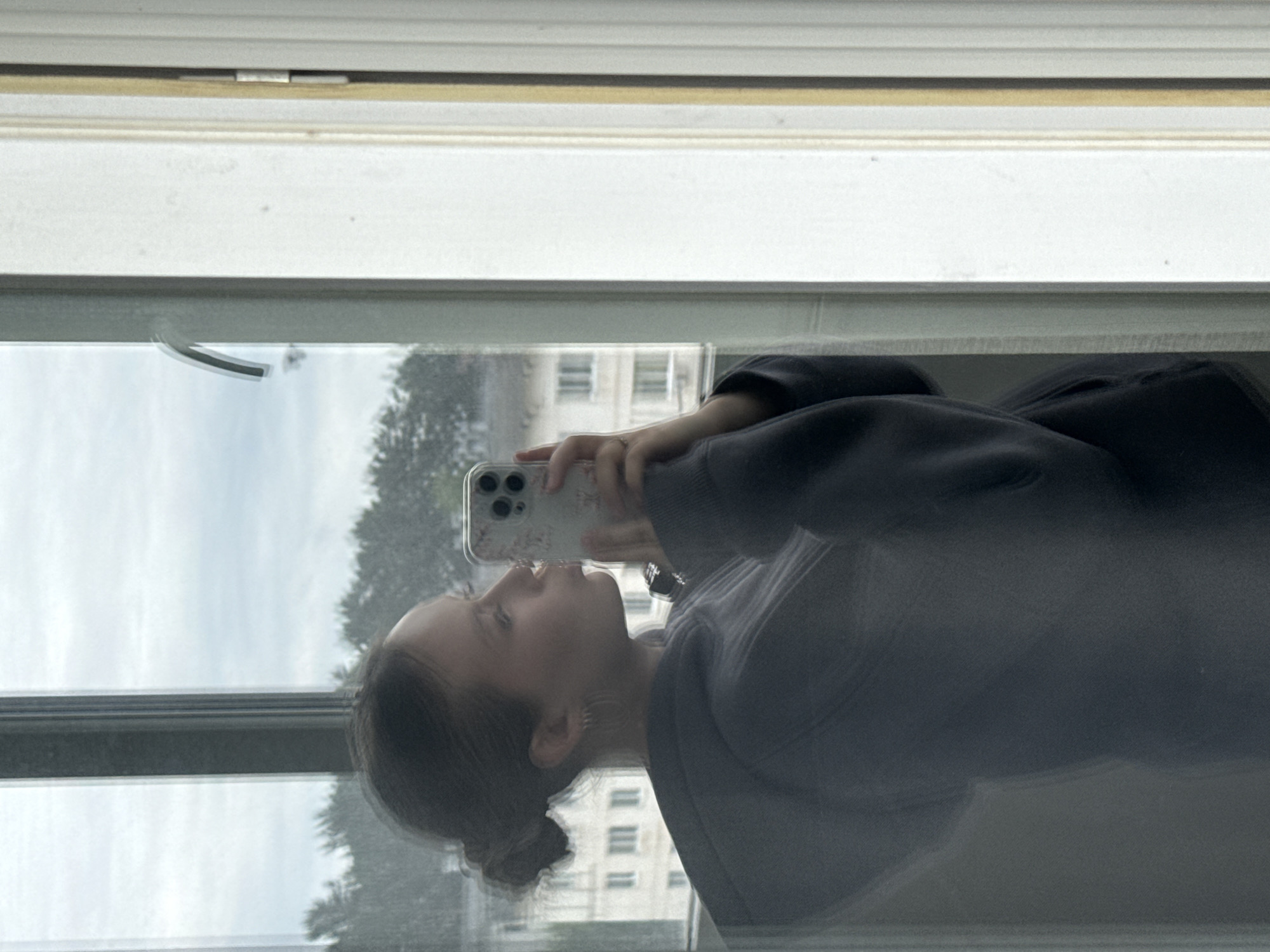 A young woman is captured in a reflection in a window, appearing to take a selfie with her smartphone. She is shown from the chest up, looking slightly upwards towards the phone, which she holds with her right hand. Her dark hair is pulled back into a bun, and she wears a dark-colored long-sleeved top or hoodie. A ring is visible on the ring finger of her right hand, and a decorative earring is on her right ear.
The smartphone, likely an iPhone due to its triple camera array, has a clear or light-colored case with a subtle floral or patterned design. The window frame, predominantly white, is visible along the top of the image (when oriented correctly), indicating she is indoors looking out. Through the window and also reflected on its surface, an overcast sky, green trees, and multi-story buildings can be seen in the distance, suggesting an urban or suburban setting. The lighting is soft and diffused, consistent with an overcast day. No explicit text is visible. The scene takes place in Chaikovsky, Russia.