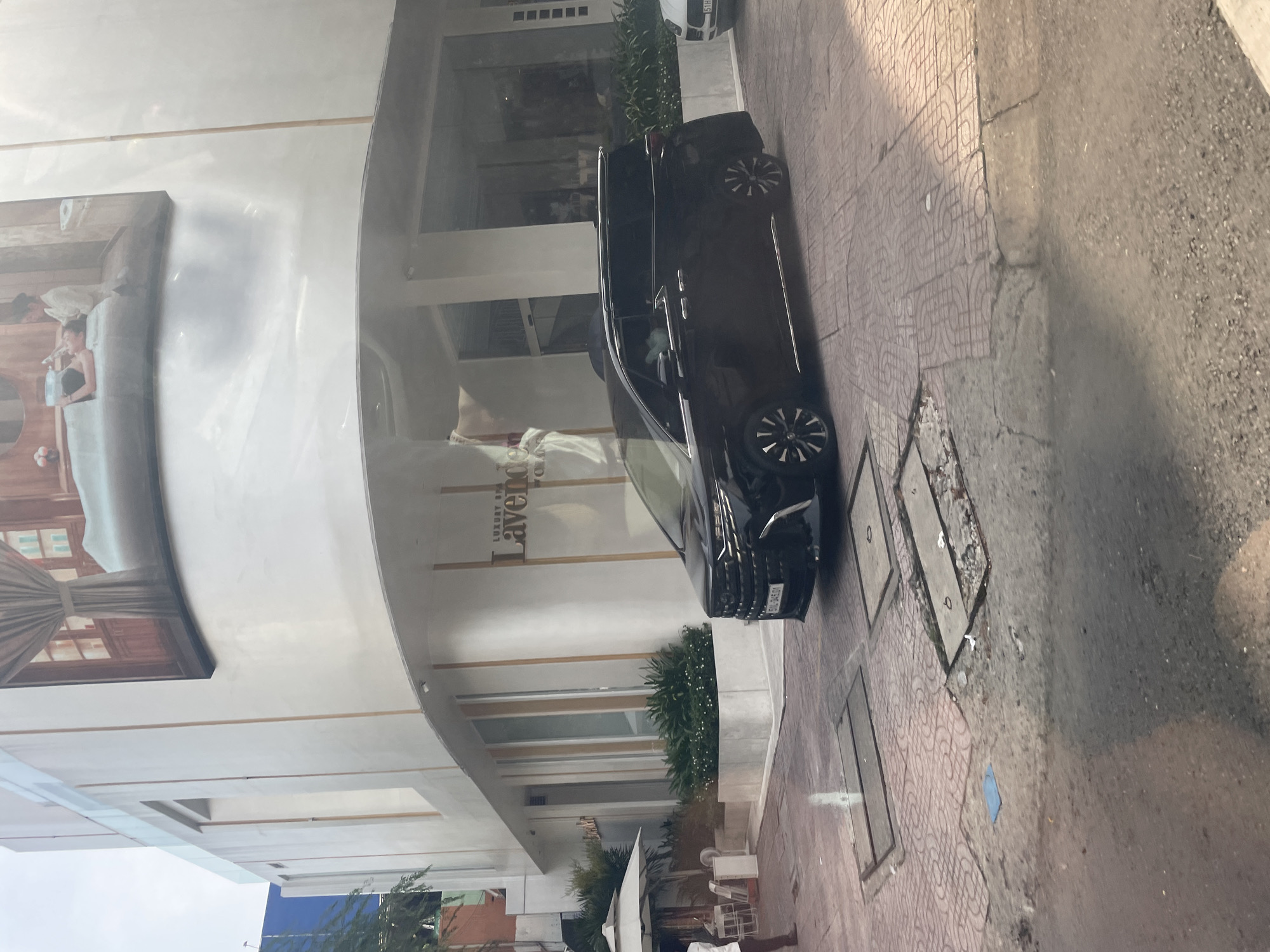 In Thủ Đức, Vietnam, a black minivan is parked on a street in front of a modern building. The building features a curved facade with a large mural depicting a spa scene on its upper level. Below the mural, the building has a white exterior with glass windows and vertical wooden accents. A sign on the building reads "LUXURY SPA Lavende".
To the left of the minivan, there is an outdoor seating area with umbrellas and tables, suggesting a cafe or restaurant. A person in a white shirt and dark pants is standing near this area. The street is paved with patterned tiles and concrete slabs. The scene is captured during daylight, with sunlight casting shadows on the ground.