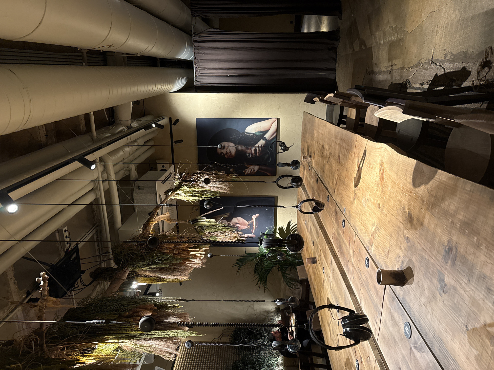 This is an interior shot of a space in Barcelona, Spain, possibly a recording studio or a music-related establishment. The image is taken from a low angle, looking upward.
The ceiling has exposed industrial pipes. Below the pipes are track lighting fixtures and what appear to be suspended decorative elements made of dried plants. Two framed art pieces hanging on the wall depict female figures. Headphones hang from the wall, attached to cables that run to the artwork.
A long wooden table occupies the right side of the frame. It's positioned vertically in the image and has cylindrical metal pieces sticking out from it. More sets of headphones are hanging along the table.