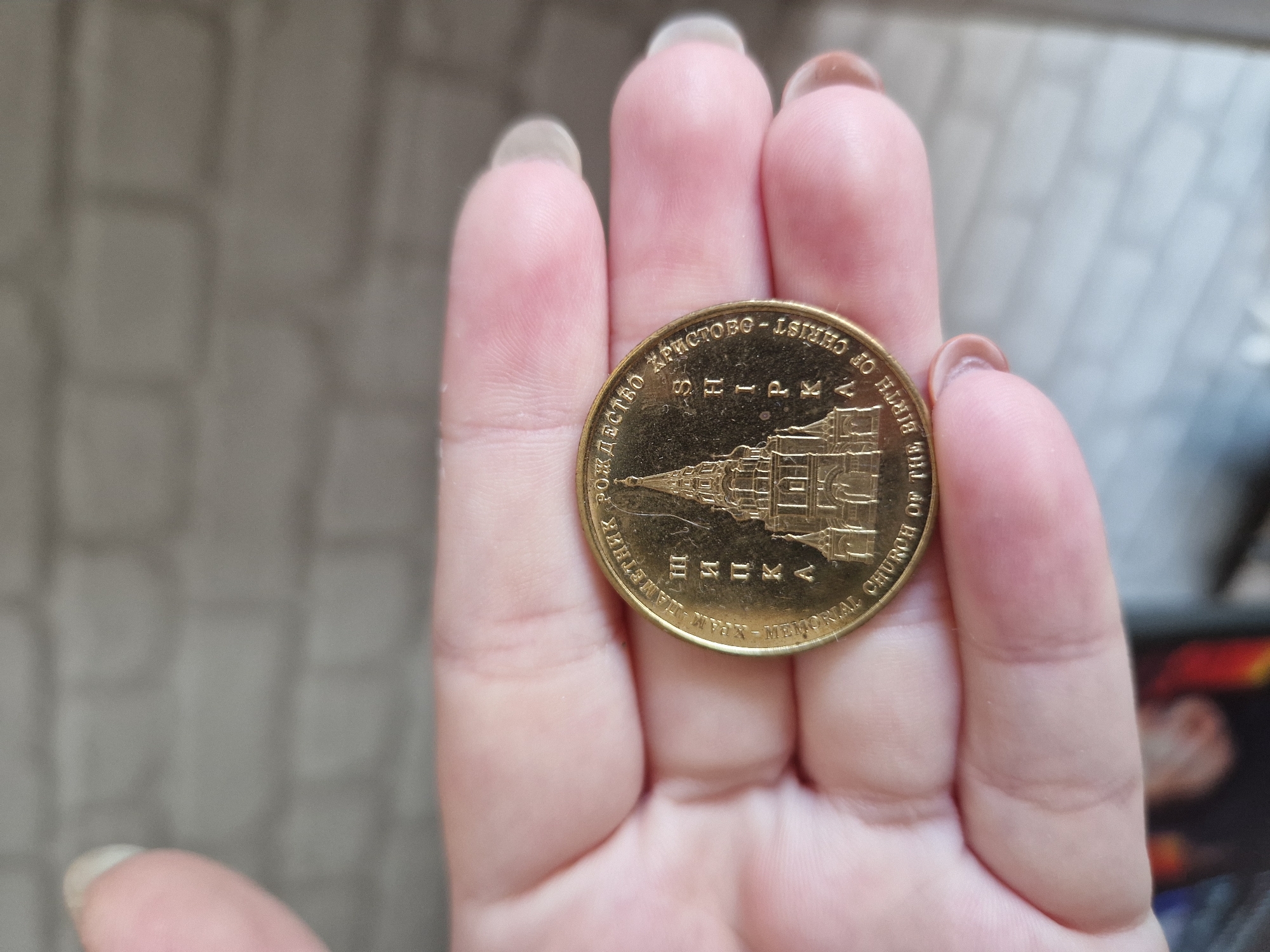 A light-skinned hand, likely a left hand, holds a gold-colored commemorative medal or coin. The medal features a detailed, raised engraving of the Shipka Memorial Church, a multi-tiered Eastern Orthodox church building with multiple spires and domes. Circular inscriptions frame the central image. The Cyrillic text "ПАМЕТНИК РОЖДЕСТВО ХРИСТОВО - ШИПКА" and the English text "MEMORIAL CHURCH OF THE BIRTH OF CHRIST - SHIPKA" are clearly legible on the medal's surface. The hand's fingers are slightly curved, securing the medal. The fingernails are light-toned and appear to be of medium length with a rounded shape. The background is an out-of-focus, light grey-brown wall with faint rectangular patterns, suggesting brickwork or tiles. The lighting is soft and even. The location depicted on the medal is Shipka, Bulgaria.