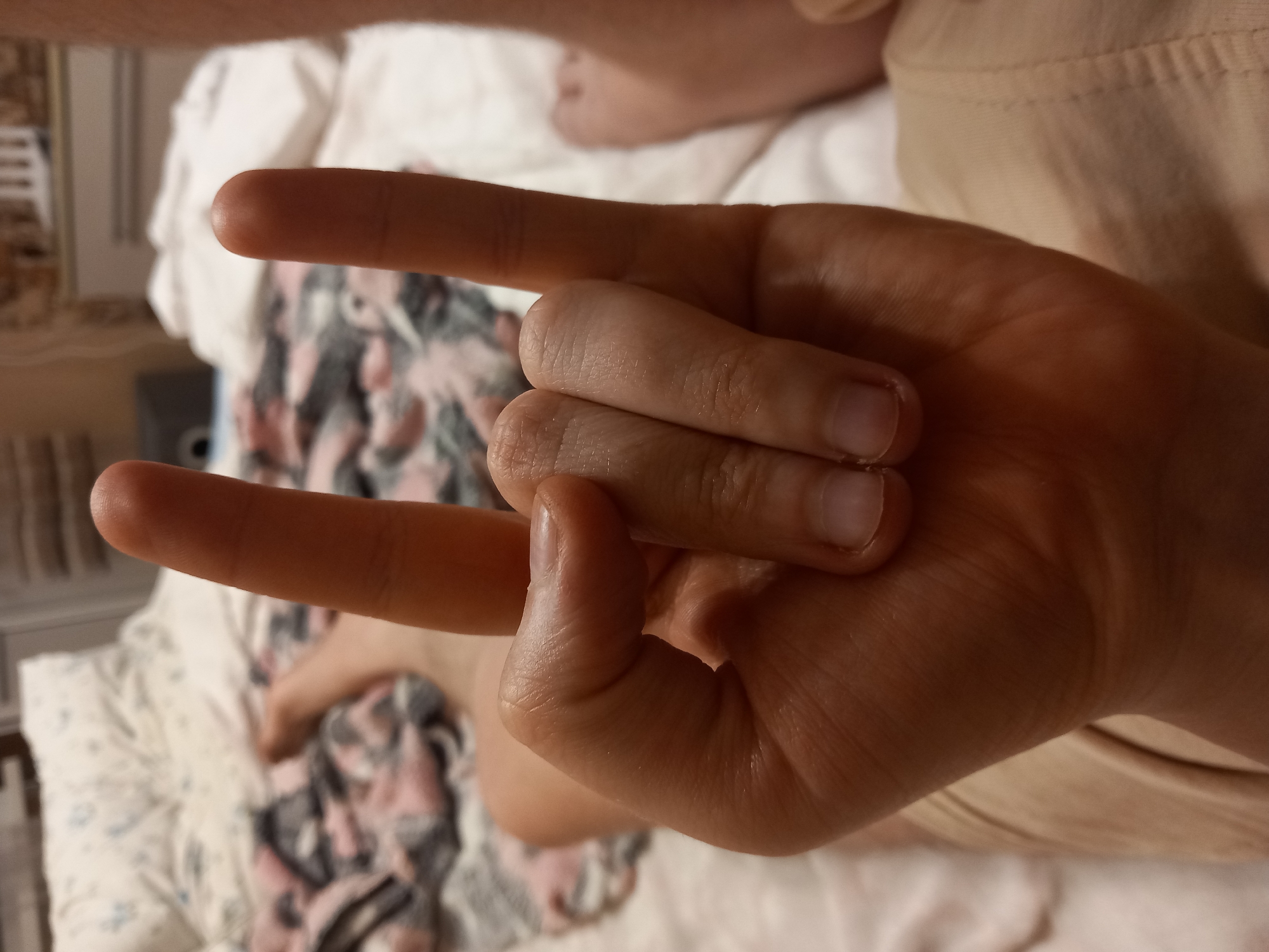 The image shows a close-up of a person's hand forming the "rock on" gesture. The hand is positioned in the foreground, with the index and pinky fingers extended upwards and apart, while the thumb, middle, and ring fingers are curled downwards. The skin on the hand appears to be light-toned. The background is blurred, but it suggests an indoor setting. There is a patterned blanket or bedspread visible behind the hand, with colors of white, pink, and gray. A sliver of a person's leg, with light skin, is also visible in the upper background. The lighting suggests it is daytime or a well-lit interior. There is no visible text in the image.