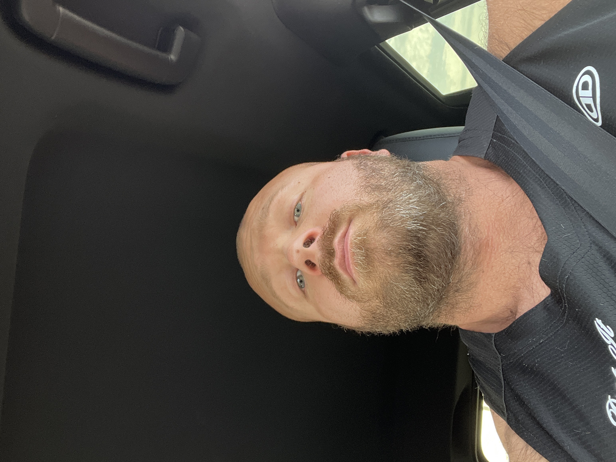 This is a close-up, low-angle, front-facing selfie taken inside a vehicle, likely a car. The subject is a bald, fair-skinned man with a full beard and light blue eyes. He is wearing a black t-shirt with a white logo on the right side. A black seatbelt crosses diagonally over his chest. The interior of the vehicle appears to be black. A window with a bright, overcast sky is visible on the right side of the frame. The exact location in Chaikovsky, Russia, cannot be determined from the image alone.
