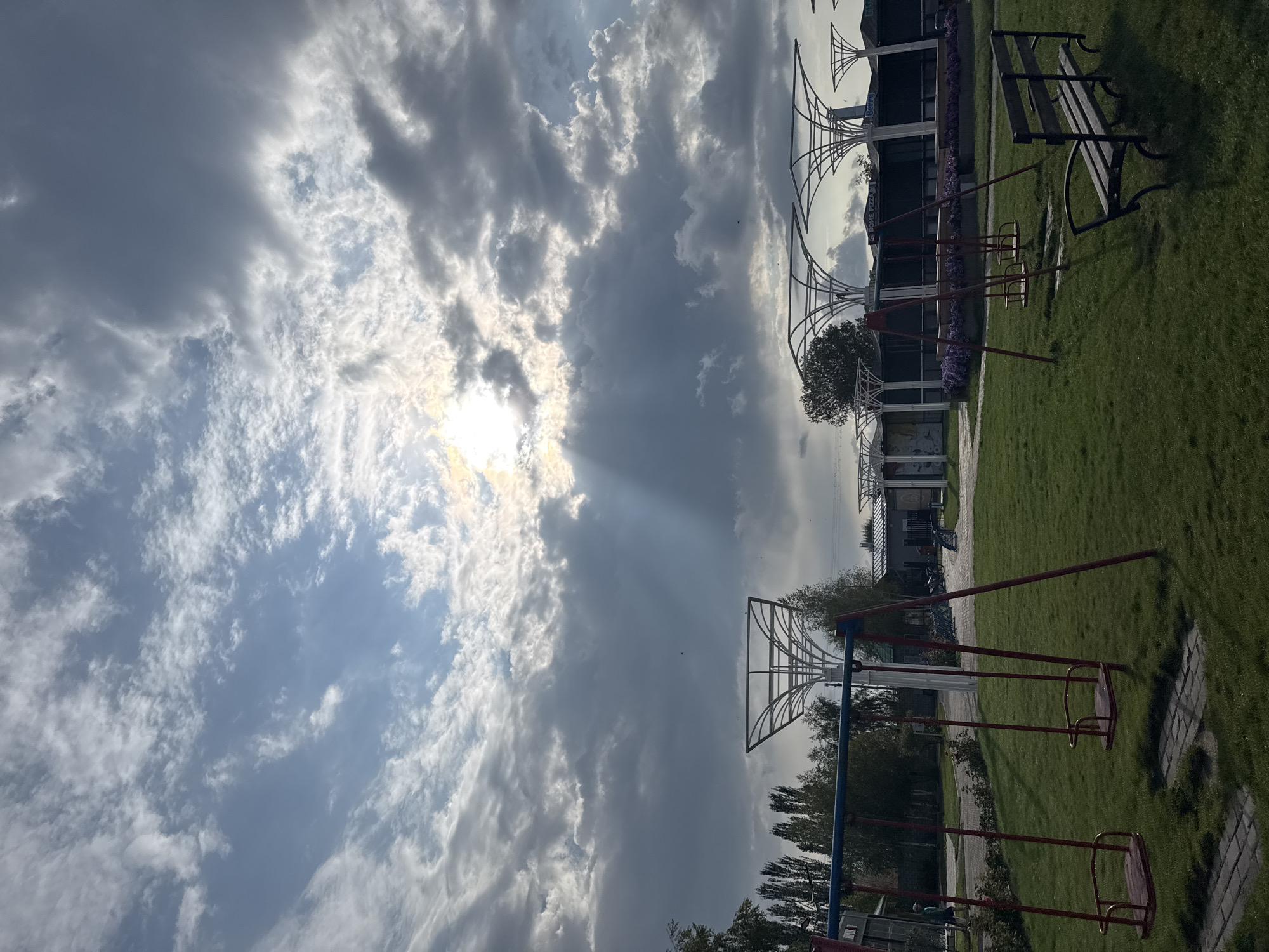 The image depicts a public park area in Sary-Oy, Kyrgyzstan, under a dramatic, cloud-filled sky with the sun breaking through in rays. In the foreground, a well-maintained grassy area features a set of red metal swings with wooden seats and a dark wooden park bench. Behind the swings, several modern, white, umbrella-like structures provide shade. Further back, trees and some buildings are visible. The overall scene suggests a tranquil, possibly late afternoon setting in a recreational space.