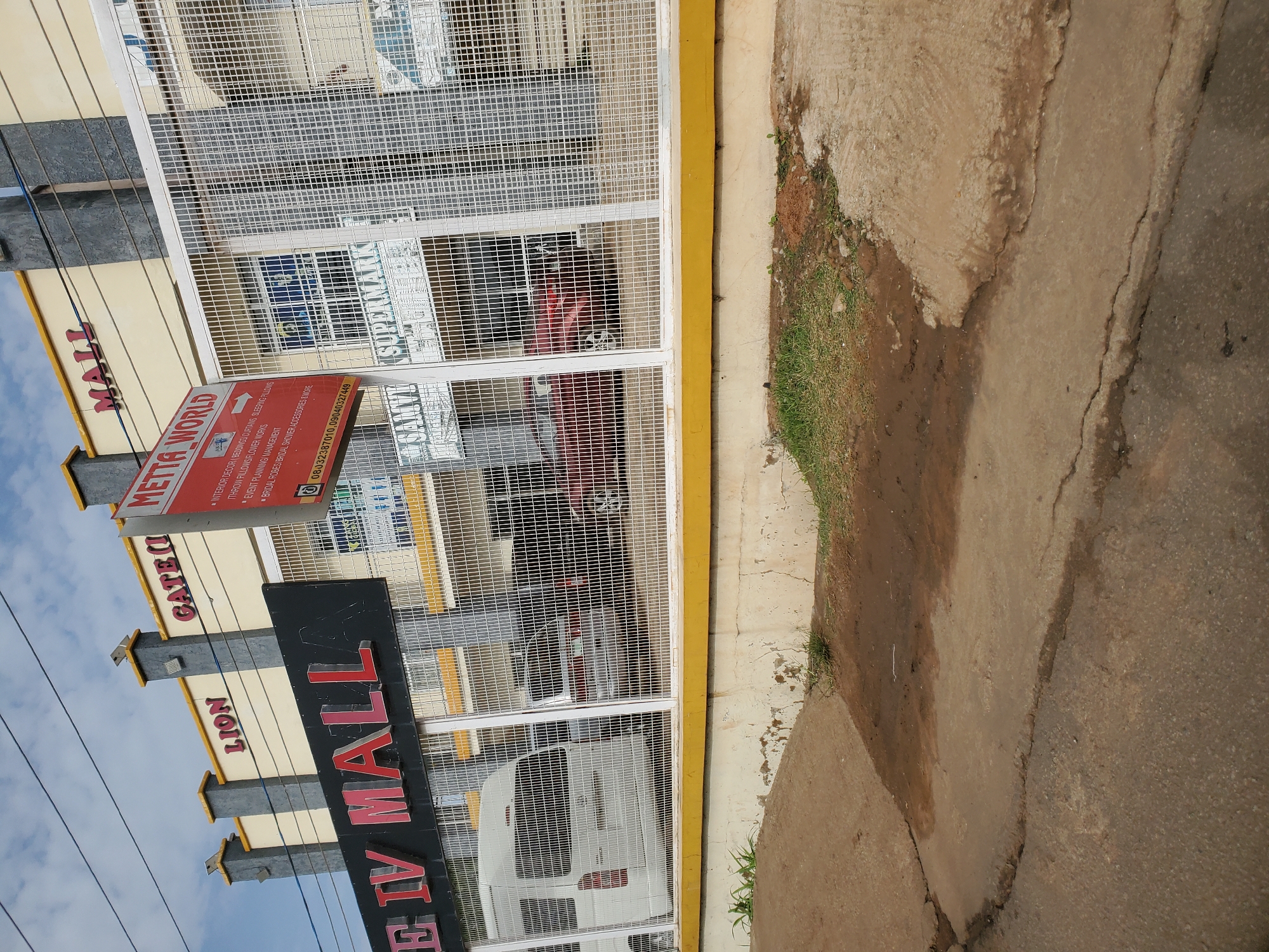 The image shows a street view of the Lion Gate IV Mall in Bukuru, Nigeria. The mall features a prominent sign with "LION GATE IV MALL" in large red letters. Above this, the words "GATE II" are visible on a building facade. Further to the right, another sign reads "METTA WORLD" and lists services such as "INTERIOR DECOR, BEDDINGS, CURTAINS, SLEEPING PILLOWS, THROW PILLOWS/FLOWER WORKS, EVENT PLANNING/MANAGEMENT, BRIDAL ROBES/BRIDAL SHOWER ACCESSORIES & MOP." Below this list are two phone numbers. The mall building has a modern facade with grey and yellow accents. A white metal fence separates the sidewalk from the mall property, where a white van and several cars are parked. The ground in front of the fence is a mixture of cracked concrete and dirt, with some patches of grass. The sky is cloudy.