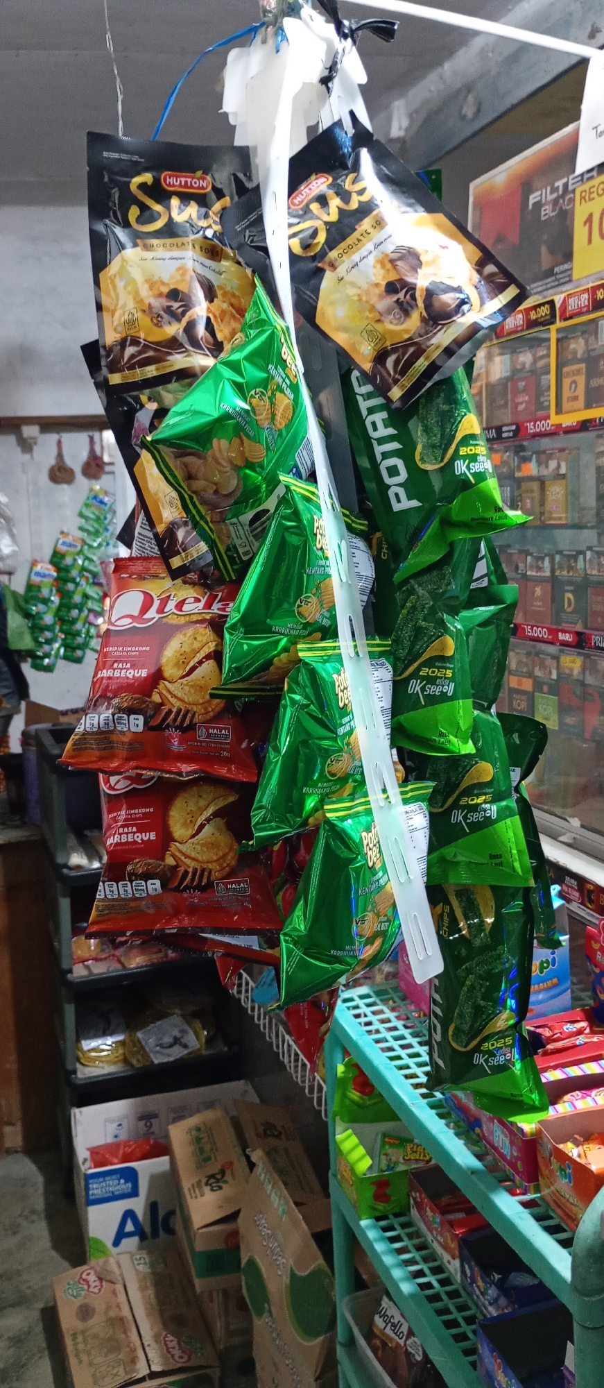 The image shows a close-up of various snack food items, primarily bags of chips and cookies, hanging from a display. The items are a mix of brands and flavors, including "Hutton Sus" chocolate snacks, "Potato Bee" chips, and "Qtela" barbecue flavored crackers. The snacks are predominantly packaged in black, yellow, and bright green bags. Some of the bags have text and logos in Indonesian, such as "HALAL" and flavor descriptions like "RASA BARBEQUE" (barbecue flavor).
The snacks are displayed on shelves and possibly hanging from the ceiling using plastic ties. In the background, there appears to be a store setting with more products on shelves, including what look like cigarette packs and other packaged goods. A portion of a blue shelf unit is visible on the right, holding more snack items. The overall environment suggests a small convenience store or a local market stall in Pendil, Indonesia, where these items are being sold. The lighting appears to be indoor, with a slightly dim atmosphere. No people are visible in the frame.