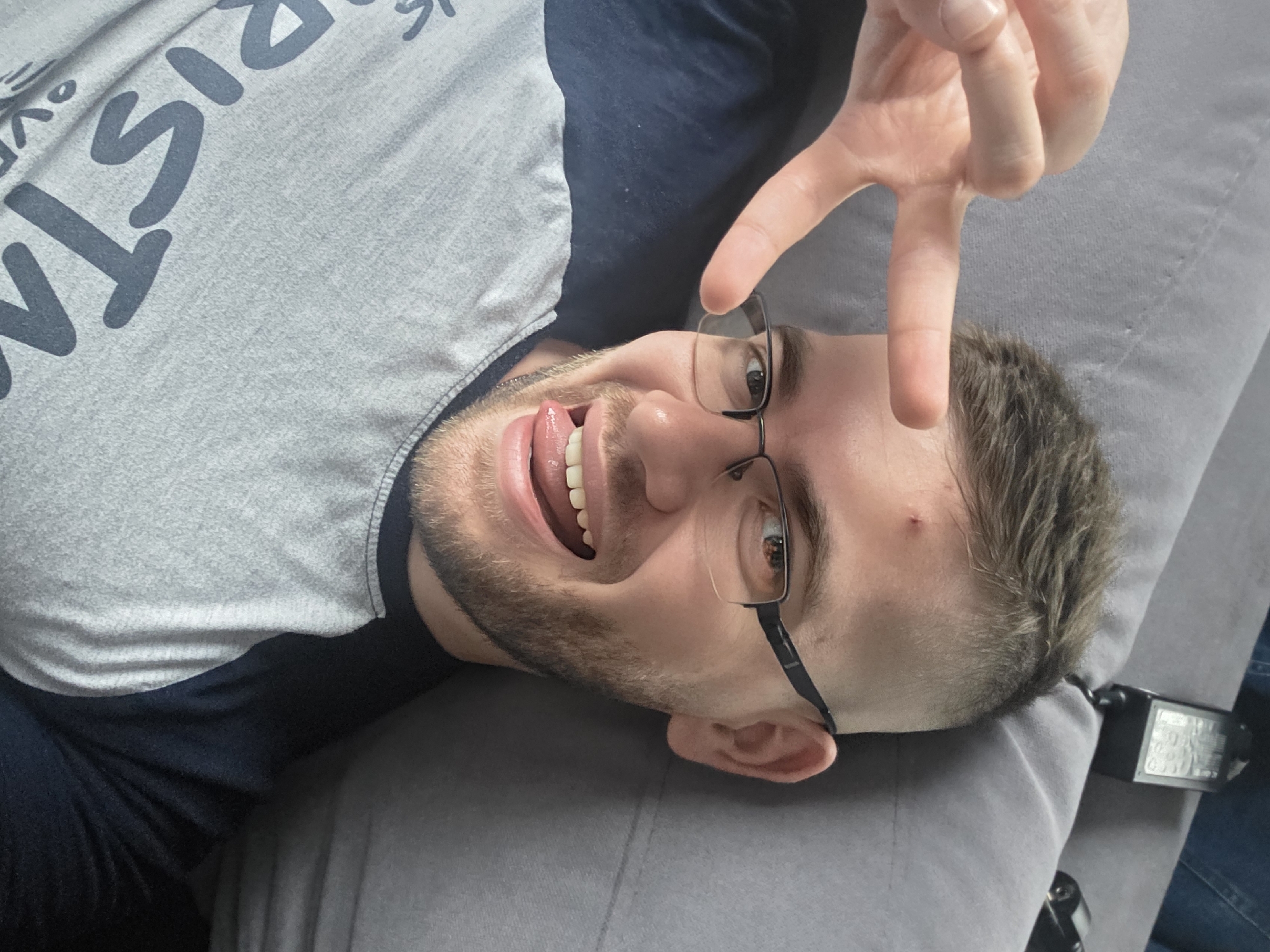 A Caucasian male, lying on a gray surface, possibly a couch or bed. He is wearing glasses and a gray t-shirt with dark sleeves. He has short brown hair and a beard. His mouth is open and his tongue is sticking out. His right hand is raised above his face, fingers forming a "V" shape. A small blemish is visible on his forehead. Location and city of Yagodovo, Bulgaria are not determinable from the image.