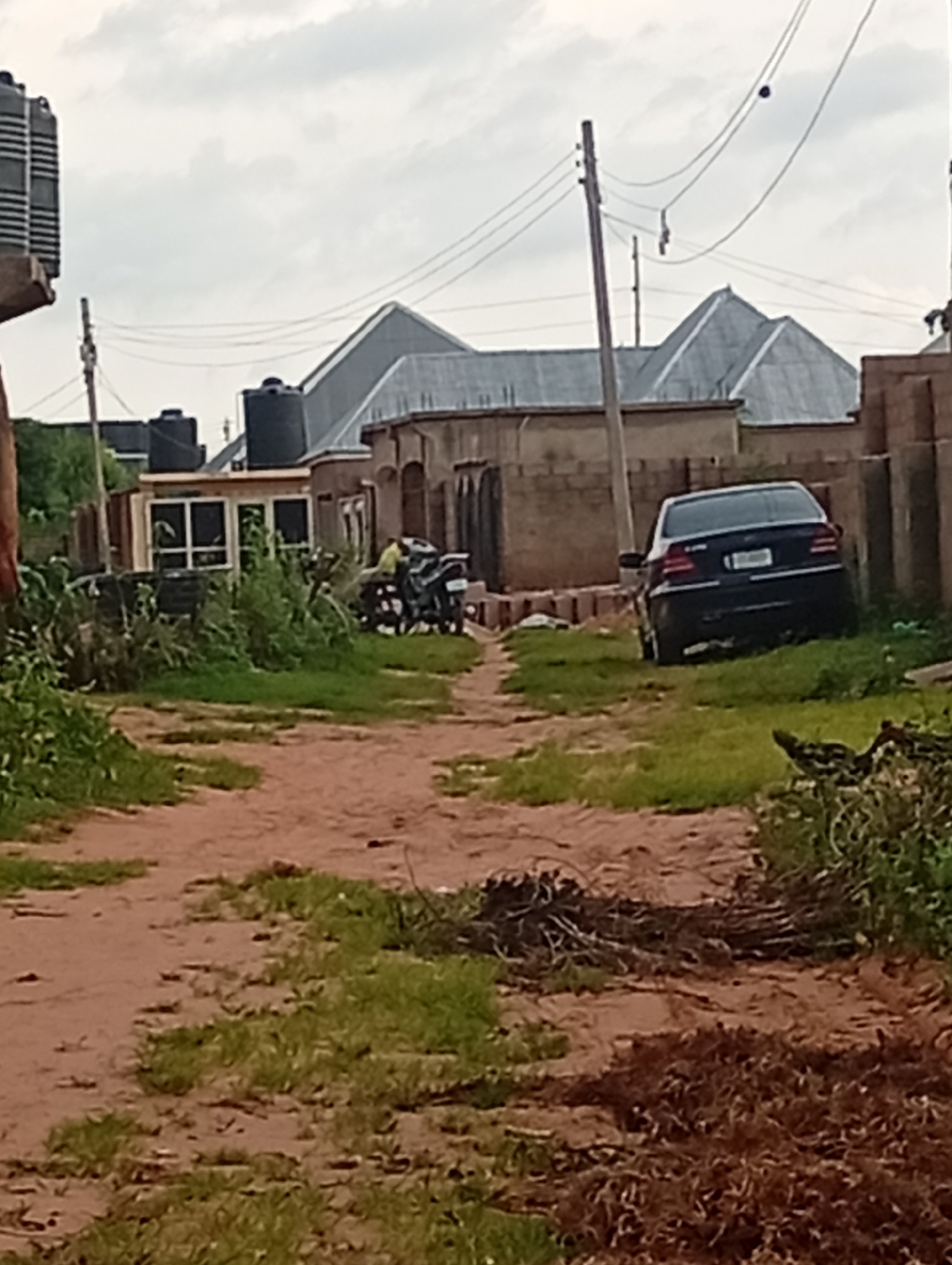 The image depicts a dirt road in Tumfure, Nigeria, lined with sparse vegetation and buildings. A black sedan is parked on the right side of the road, partially on the grass. On the left side of the road, a motorcycle with two individuals is visible. The background shows several buildings with corrugated metal roofs and water tanks. The sky is overcast with clouds.