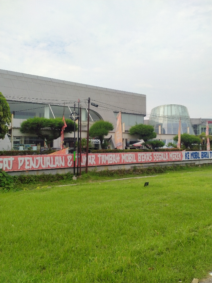 The image displays an outdoor scene in Jaten, Indonesia, under an overcast sky. In the foreground, a lush green grassy area occupies the lower half of the frame. Above this, a low concrete wall or barrier is adorned with a long banner featuring red text on a white background.
Behind the barrier, several buildings are visible. The primary structure is a modern, multi-story building with a grey facade, a flat roof, and expansive glass windows, some of which are arched. To the right, another building features a distinctive glass dome, suggesting a unique architectural element. In front of these buildings, several small, round-shaped trees are planted. Utility poles with power lines are present in the mid-ground, indicating typical urban infrastructure.
No people are visible in the scene. The lighting suggests it is daytime, with the cloud cover creating diffused light. The most prominent visible text is on the banner, which reads: "PUSAT PENJUALAN & JUAL TAMBAH MOBIL BEKAS SEGALA MEREK KE MOBIL BARU". This translates to "SALES CENTER & TRADE-IN USED CARS OF ALL BRANDS FOR NEW CARS," indicating the location likely serves as a car dealership or a car sales and exchange center. Additionally, partial text "SMOL GALLE" and a "TOYOTA" logo can be discerned on the buildings from closer inspection of the provided crops, further supporting the car dealership context.