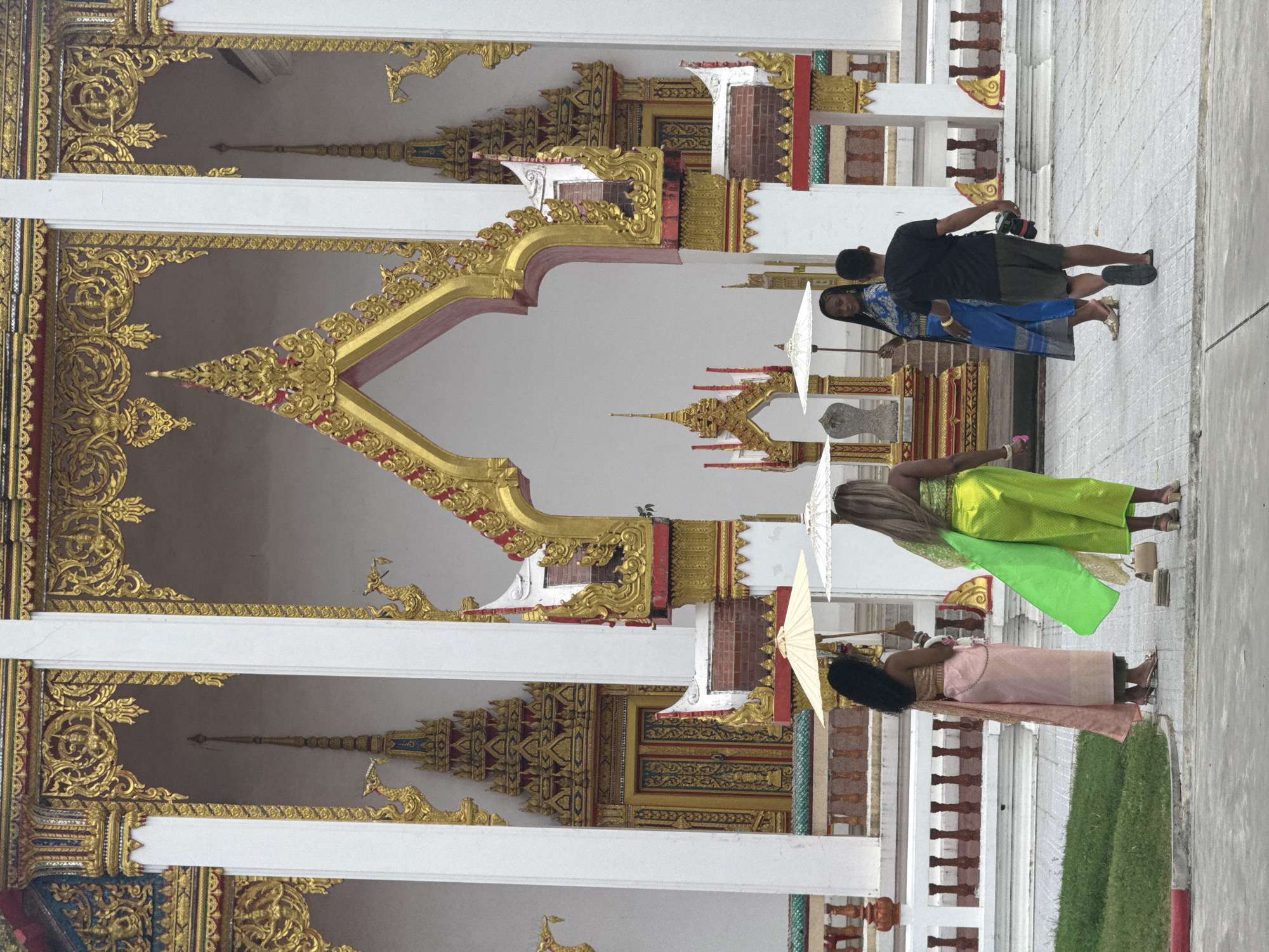 In Chalong, Thailand, at a Buddhist temple, four individuals are captured. Two women in traditional Thai attire, one in a pink skirt and golden top holding a parasol, and another in a bright green skirt and top, also holding a parasol, stand in the foreground. To their right, a third woman in a blue traditional outfit holds a parasol, facing the viewer. A man with dark hair, wearing a black t-shirt and dark shorts, walks away from the camera, carrying a camera. The scene is set against the backdrop of an ornate temple with golden decorations and white pillars.