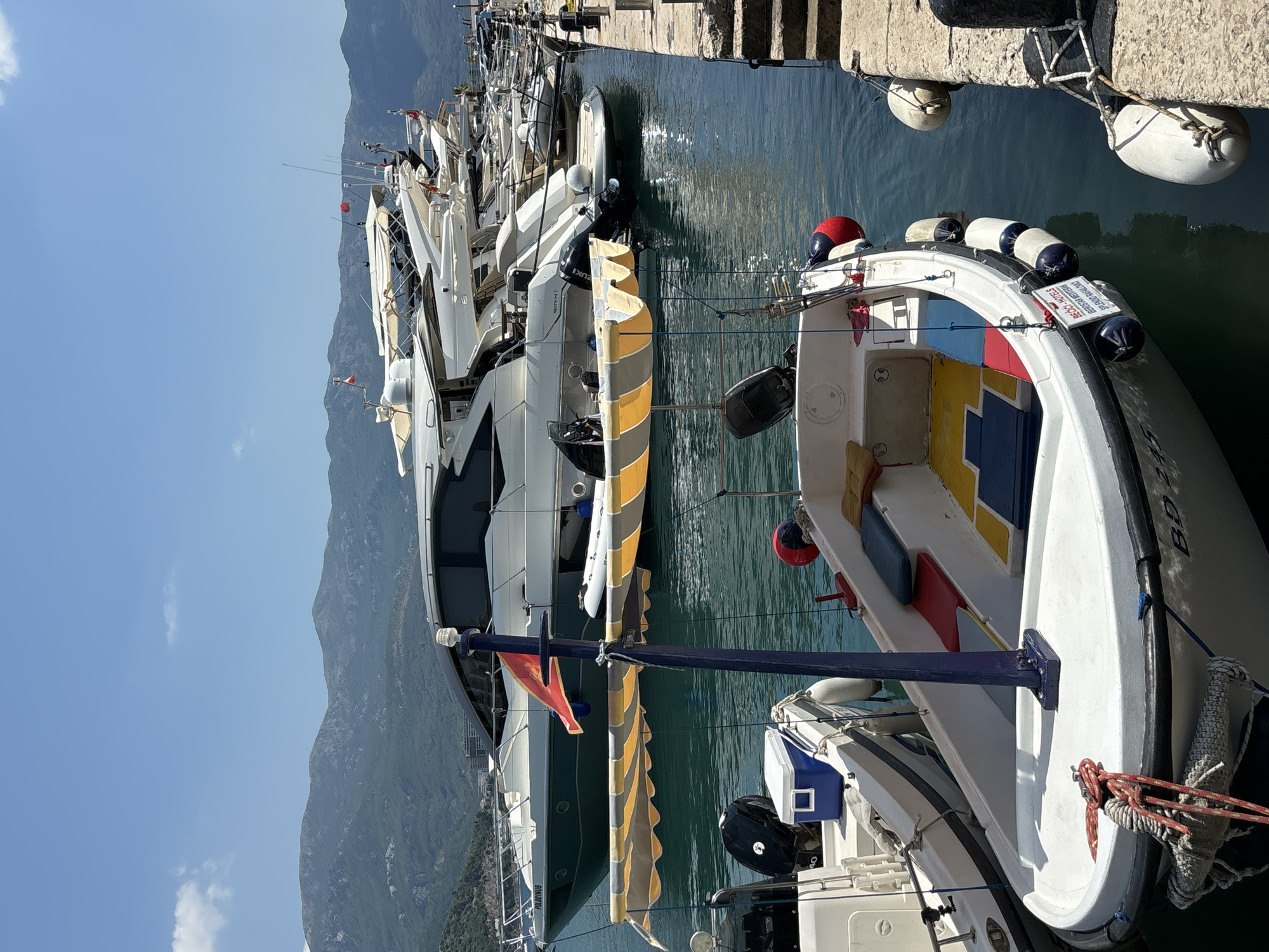 The image depicts a marina in Budva, Montenegro, with several boats docked. The scene is set on a sunny day, with clear blue skies and distant mountains. The water is calm and reflects the sunlight.
The primary subjects are various boats, ranging from large yachts to smaller motorboats. One prominent object is a white motorboat in the foreground, adorned with colorful cushions and a blue mast. Several white and blue fenders are attached to the boats and the dock. A red and blue cap rests on the bow of the foreground boat.
The boats are moored along a stone dock. In the background, a line of larger white yachts is visible, some with yellow and white awnings. A Montenegrin flag is attached to one of the masts.
The time of day appears to be late morning or early afternoon, given the bright sunlight and shadows. The weather is fair and warm, indicated by the clear sky. The location is identifiable by the Montenegrin flag and the general scenery typical of a coastal European marina.
Visible text on the foreground boat reads "BEACH HOTELS" and "SPORT". Another boat in the background has text that appears to be "POLIDOR 1909".