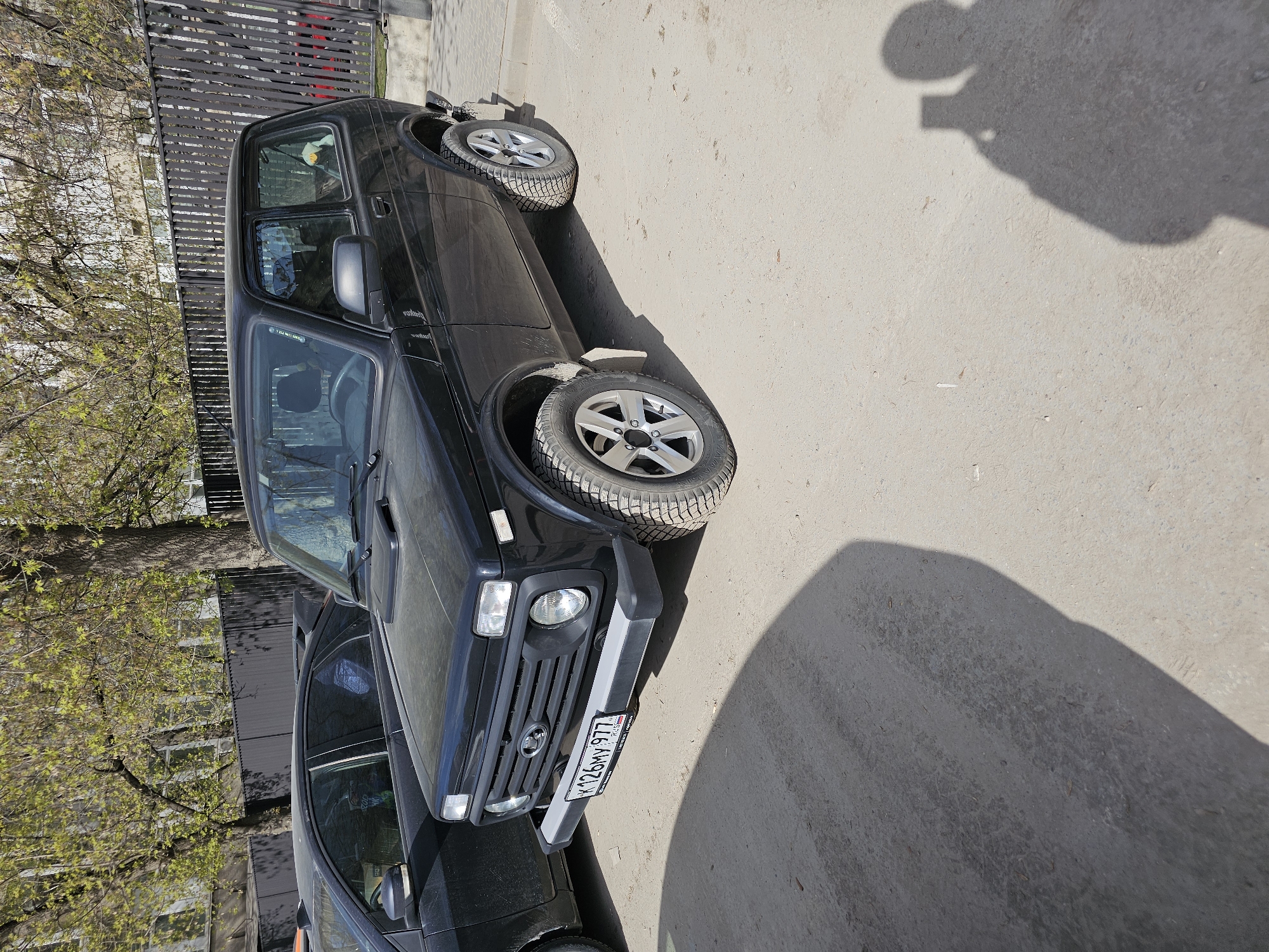 A black Lada Niva SUV is parked on a paved surface alongside a dark-colored sedan in an urban environment. The Niva's license plate, visible on the front bumper, reads "K 126 MY 977 RUS". The paved surface is light gray and exhibits some minor cracking. In the background, a metal fence runs parallel to the vehicles, with trees with budding leaves visible beyond. The shadow of the person taking the photograph is in the lower right corner. The setting is likely Moscow, Russia, based on the license plate format.