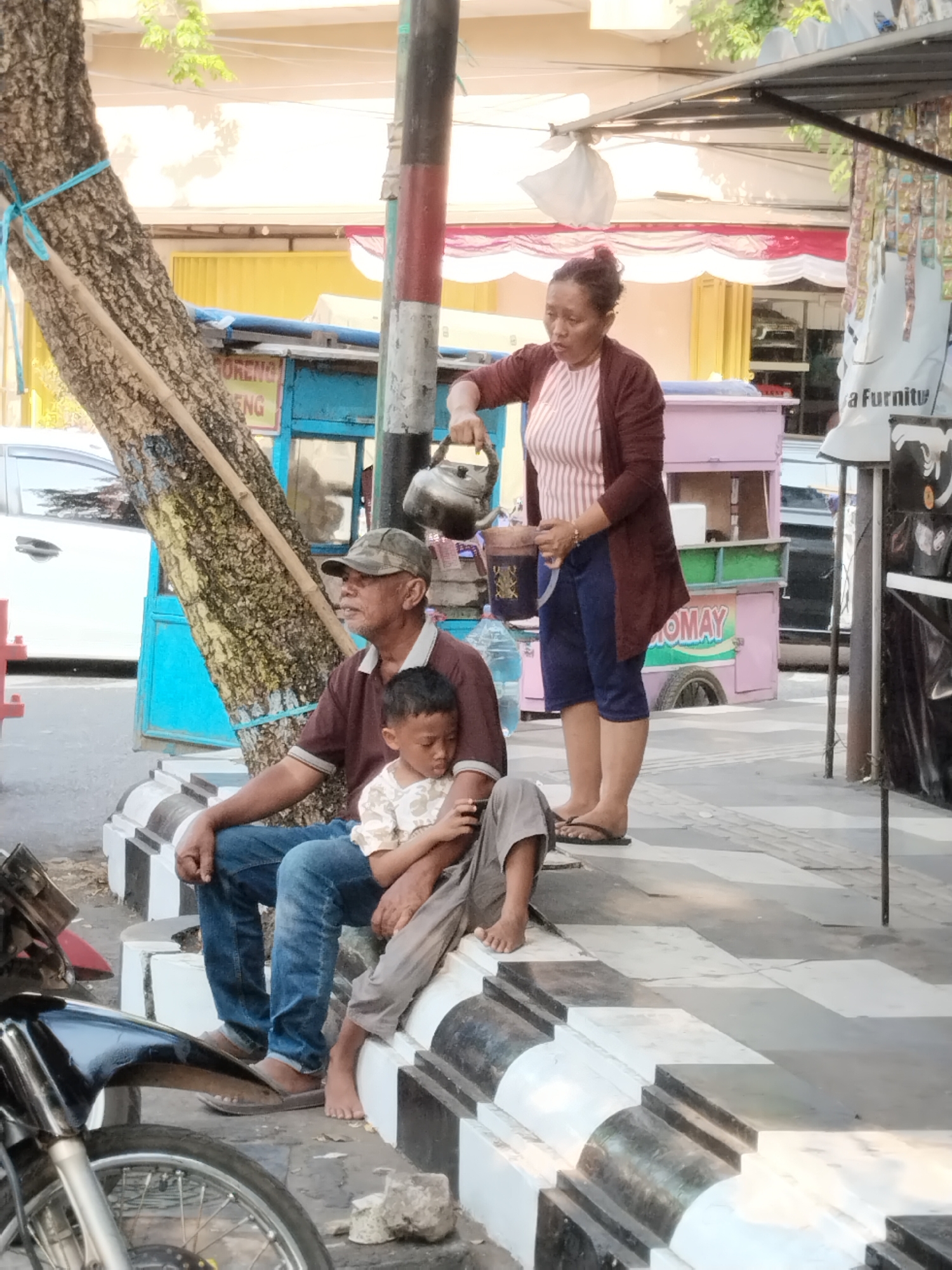 In Kecamatan Pati, Indonesia, an adult woman with dark hair tied back is pouring liquid from a metal teapot into a cup held by a young boy. The boy is seated with his legs crossed, wearing a light-colored shirt and gray pants. Beside him, an adult man wearing a cap, a dark maroon shirt, and blue jeans is looking to his right. The three are sitting on a tiered concrete structure with black and white checkered patterns. In the background, there are food carts, one pink and one blue, and a tree with a bamboo pole propped against it. A motorcycle is visible in the foreground. The scene appears to be a street or sidewalk area with shops or stalls.
