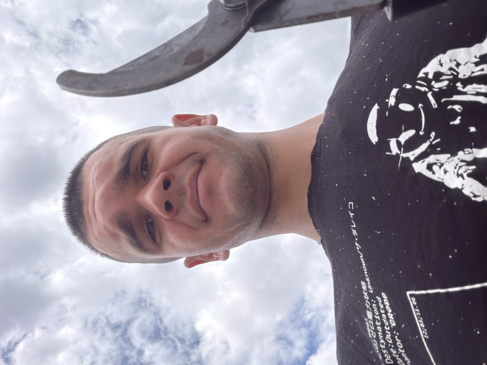 A close-up, low-angle, selfie-style shot shows a young man from the chest up, holding a pair of large, metal shears in his right hand, which is positioned to the right of the frame. The man has a buzz cut and is looking directly at the camera with a neutral expression. He is wearing a black t-shirt with white text and a graphic print of a cartoonish figure. The background is a cloudy sky. The scene appears to be outdoors, possibly in a rural or park-like setting given the presence of shears and the sky backdrop, but no specific urban features of Stryama, Bulgaria are identifiable within the image itself.