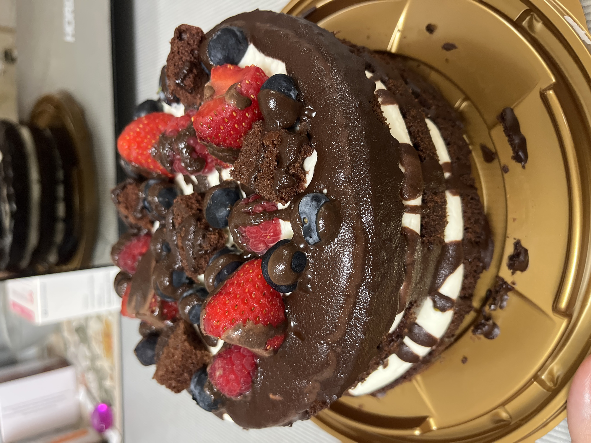A two-tiered chocolate cake is positioned on a gold plastic cake stand. The cake is decorated with dollops of white cream, chocolate ganache drizzle, fresh strawberries, raspberries, blueberries, and small pieces of brownie. The cake is placed on a white tablecloth. In the background, a blurred image of a kitchen appliance and what appears to be a box or container can be seen. The image is taken from a slightly elevated angle, focusing on the cake. The location of this scene is not specified in the image.